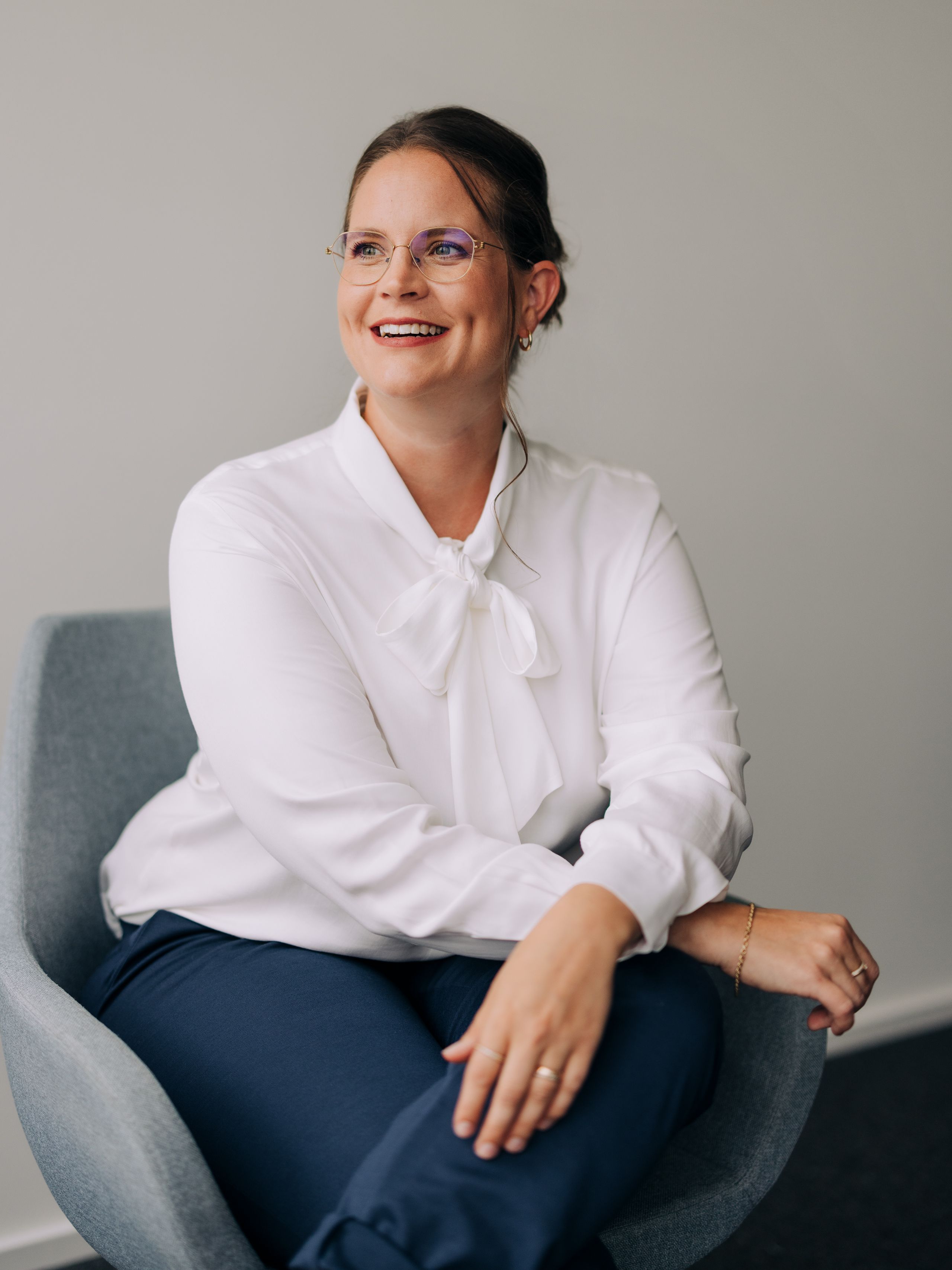 Lächelnde Frau mit Brille, weißer Bluse und blauer Hose sitzt auf einem grauen Stuhl vor einem einfarbigen Hintergrund.