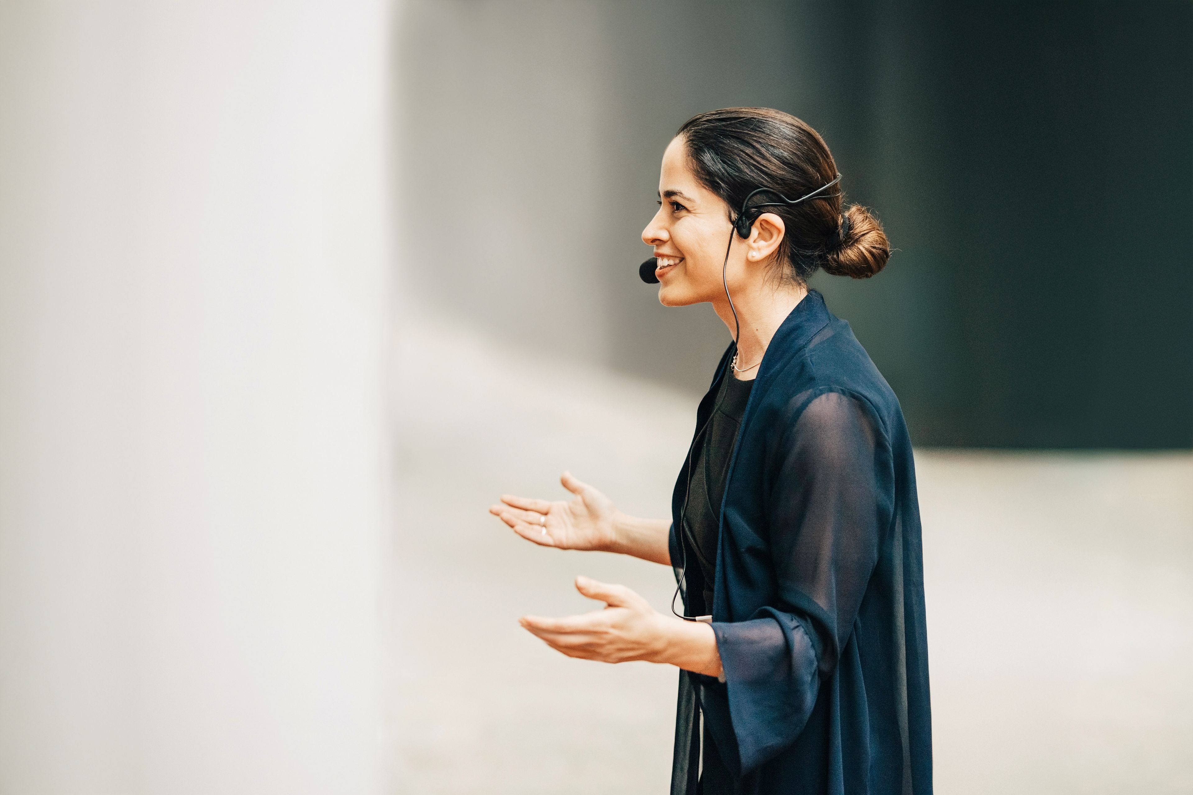 Eine Frau in dunkler Kleidung, die ein Headset trägt, gestikuliert, während sie vor einem hellen, verschwommenen Hintergrund spricht.