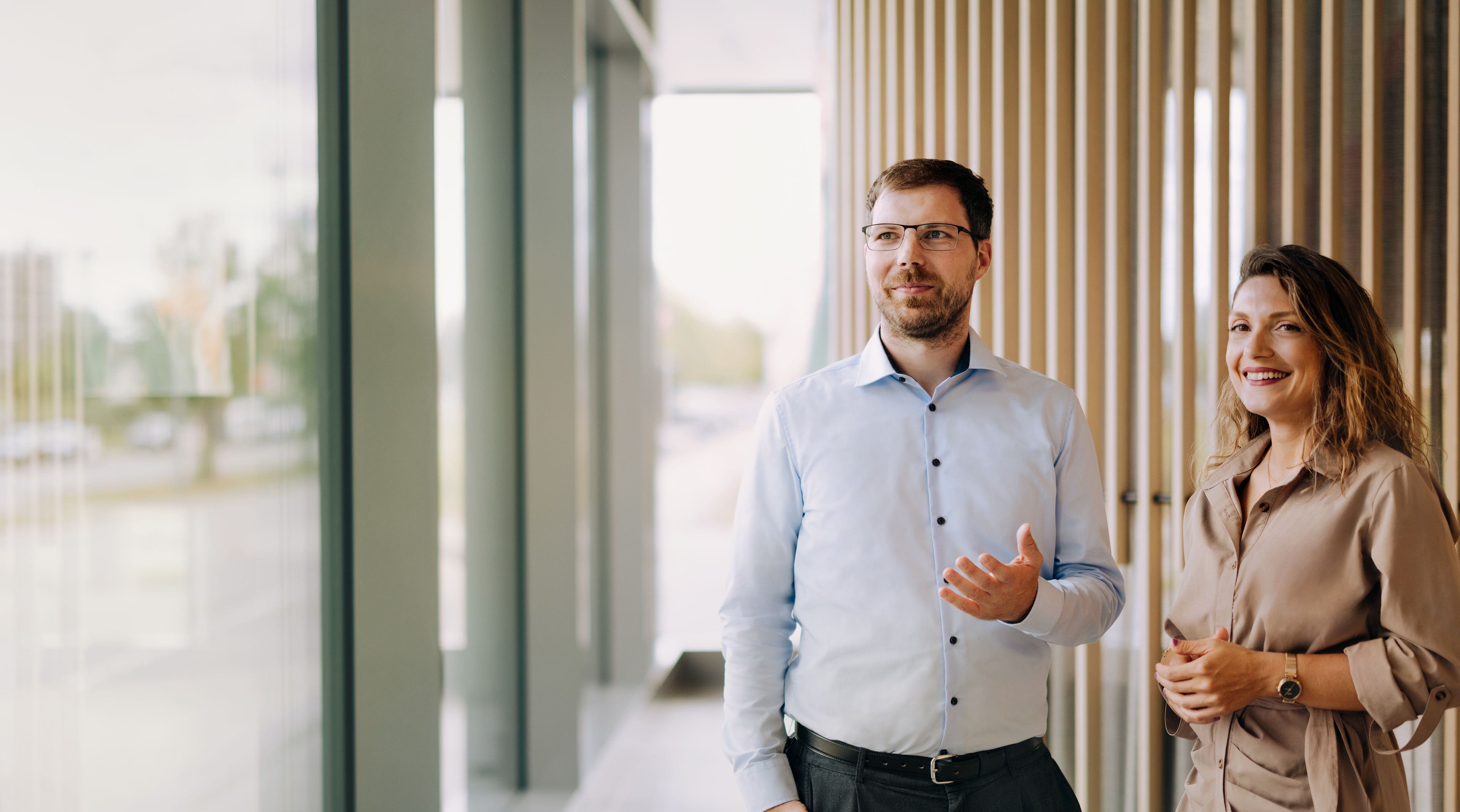 Zwei Personen stehen drinnen in der Nähe großer Fenster und unterhalten sich. Der Mann gestikuliert, und beide lächeln.