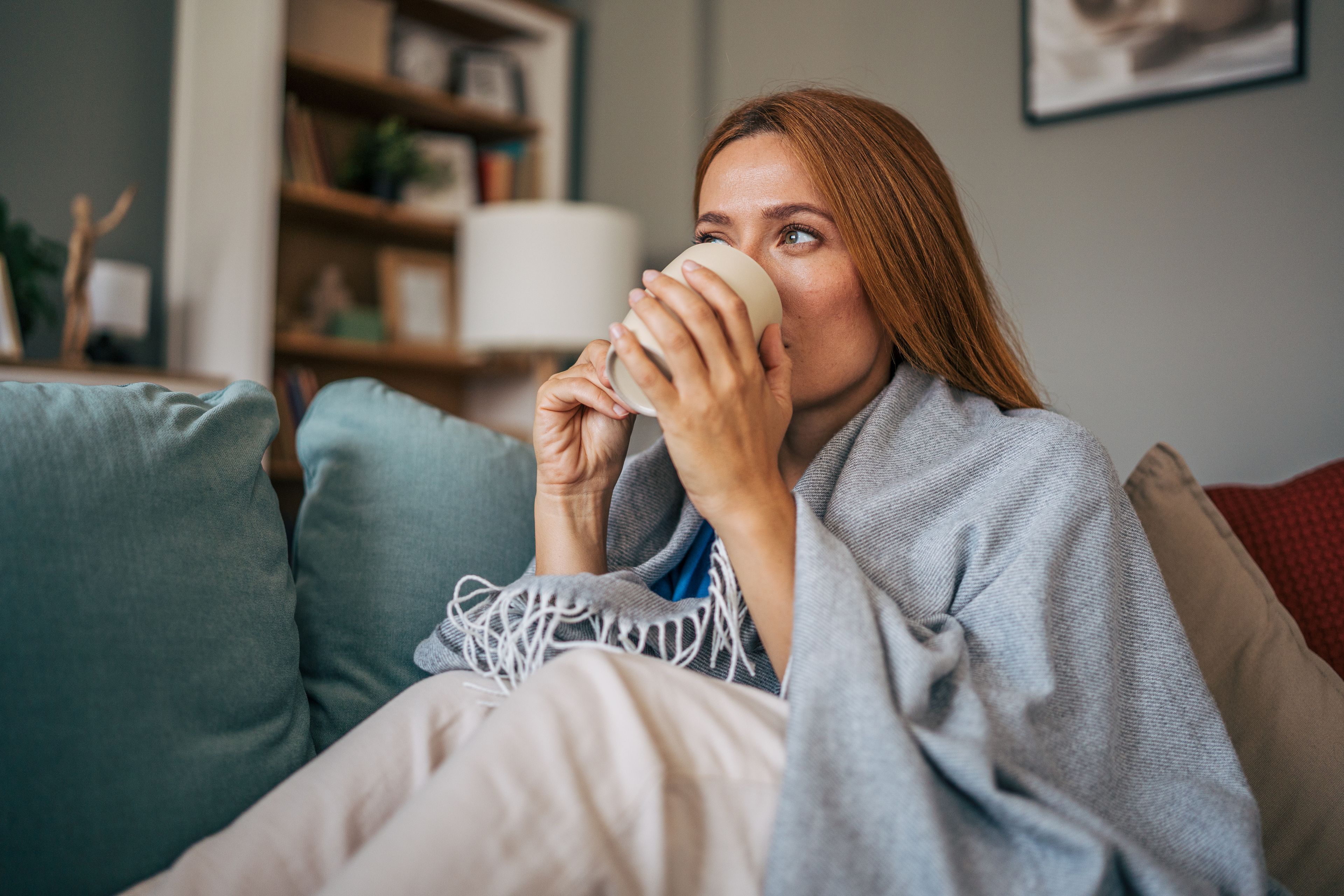 Eine Frau, die in einem grauen Deckenmantel auf einem Couch sitzt, einen Kaffee aus einem Becher in der Hand, mit Bücherregalen im Hintergrund.