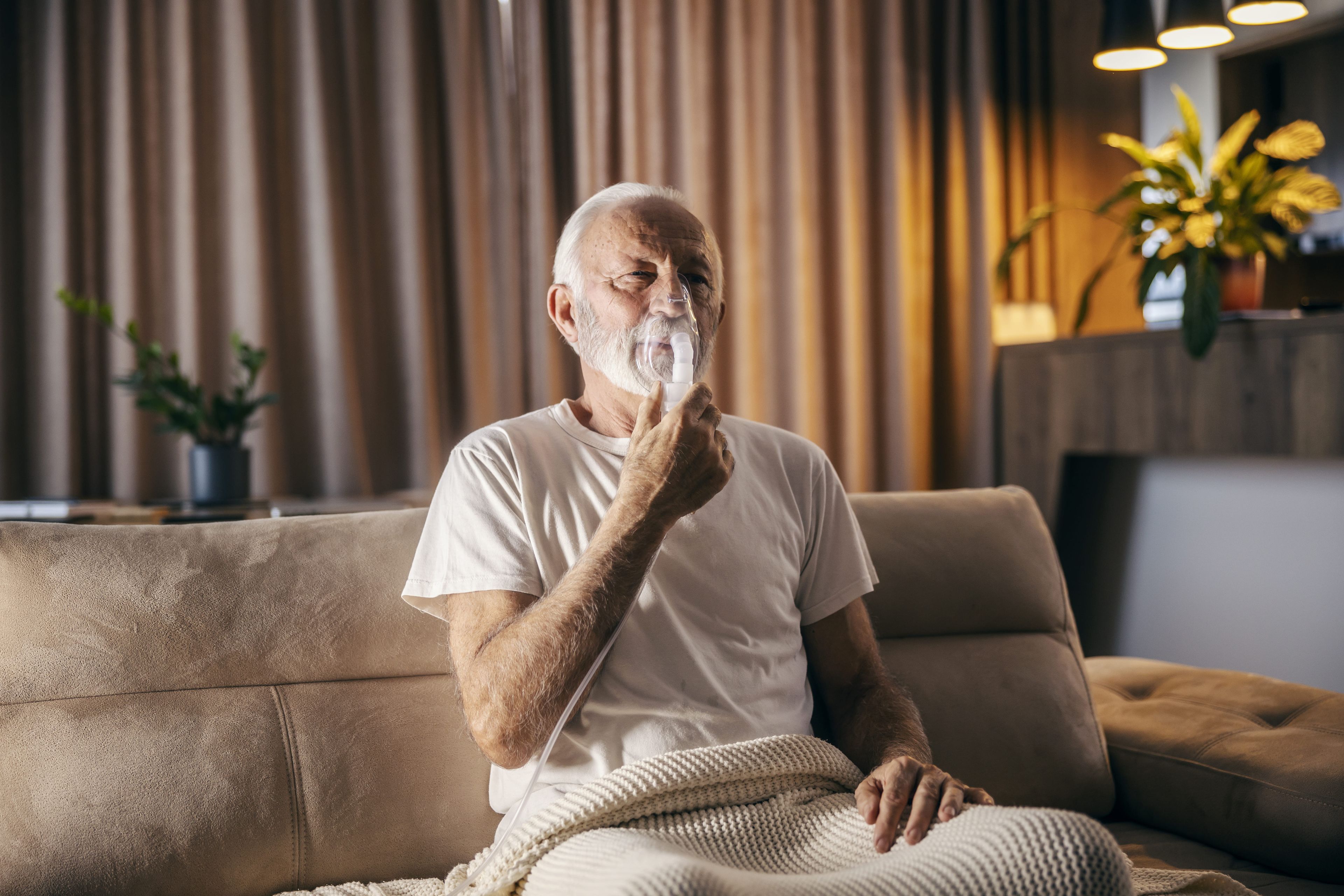 Ein älterer Mann sitzt auf einem Sofa und benutzt ein Verneblungsgerät, eingehüllt in eine Decke, in einem gemütlichen Wohnzimmer mit Pflanzen und warmer Beleuchtung.