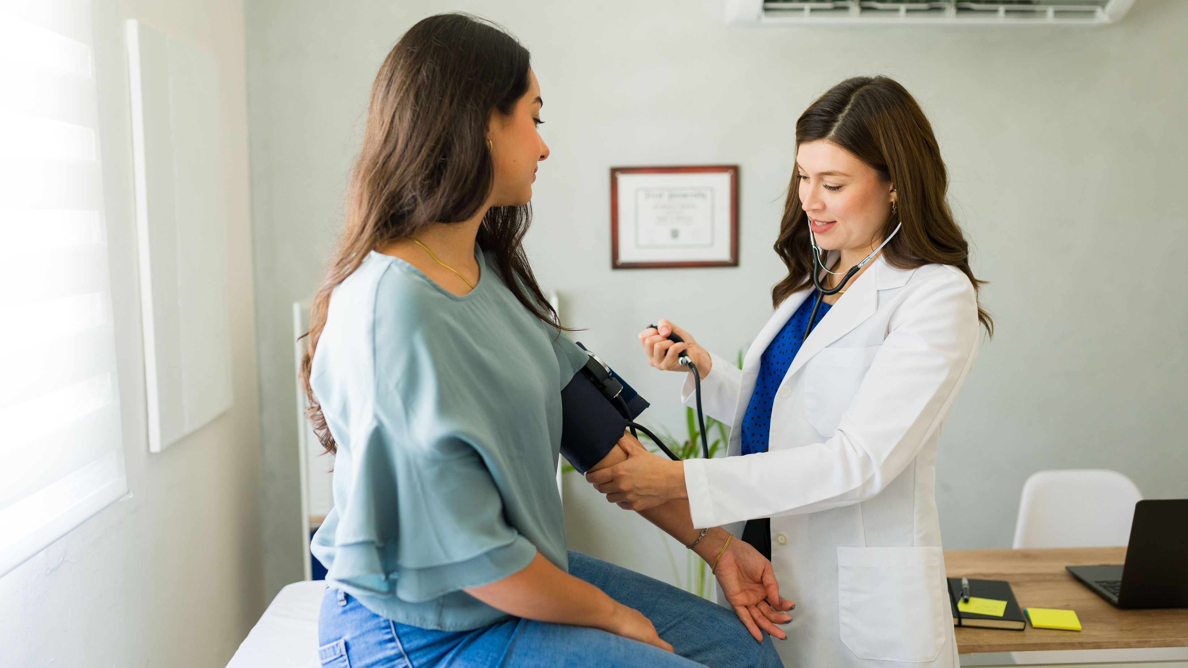 Ein Arzt misst den Blutdruck eines Patienten in einer Arztpraxis. Der Patient sitzt auf einer Untersuchungsliege, und an der Wand ist ein Diplom sichtbar.