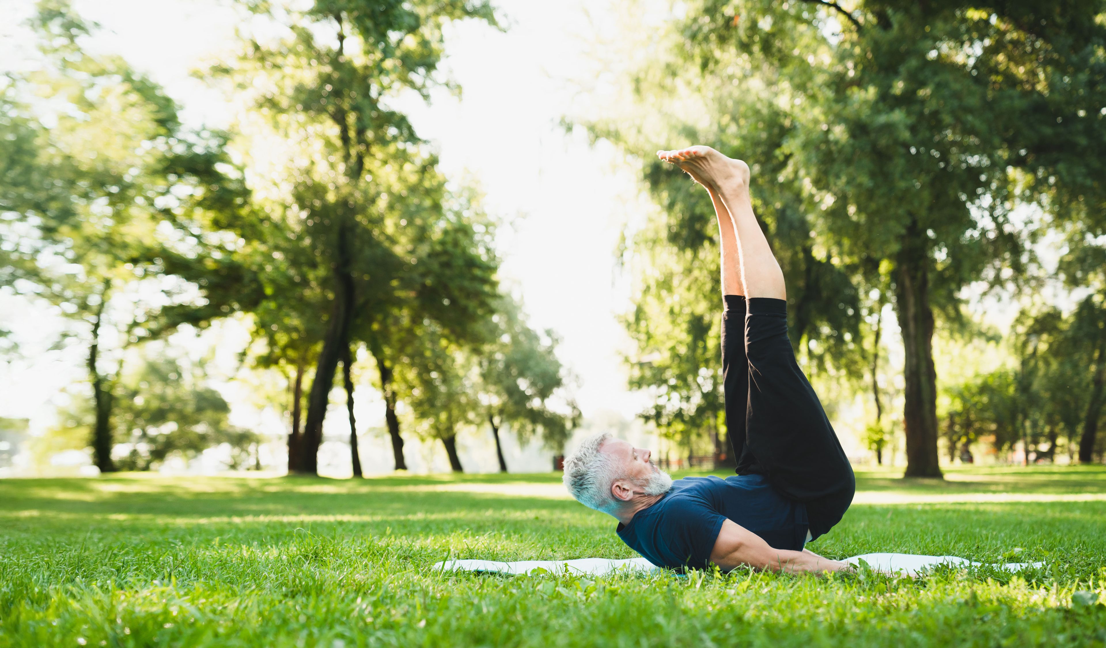 Eine ältere Person praktiziert Yoga auf einer Matte im Park und macht eine Schulterstand-Übung, umgeben von üppig grünen Bäumen.