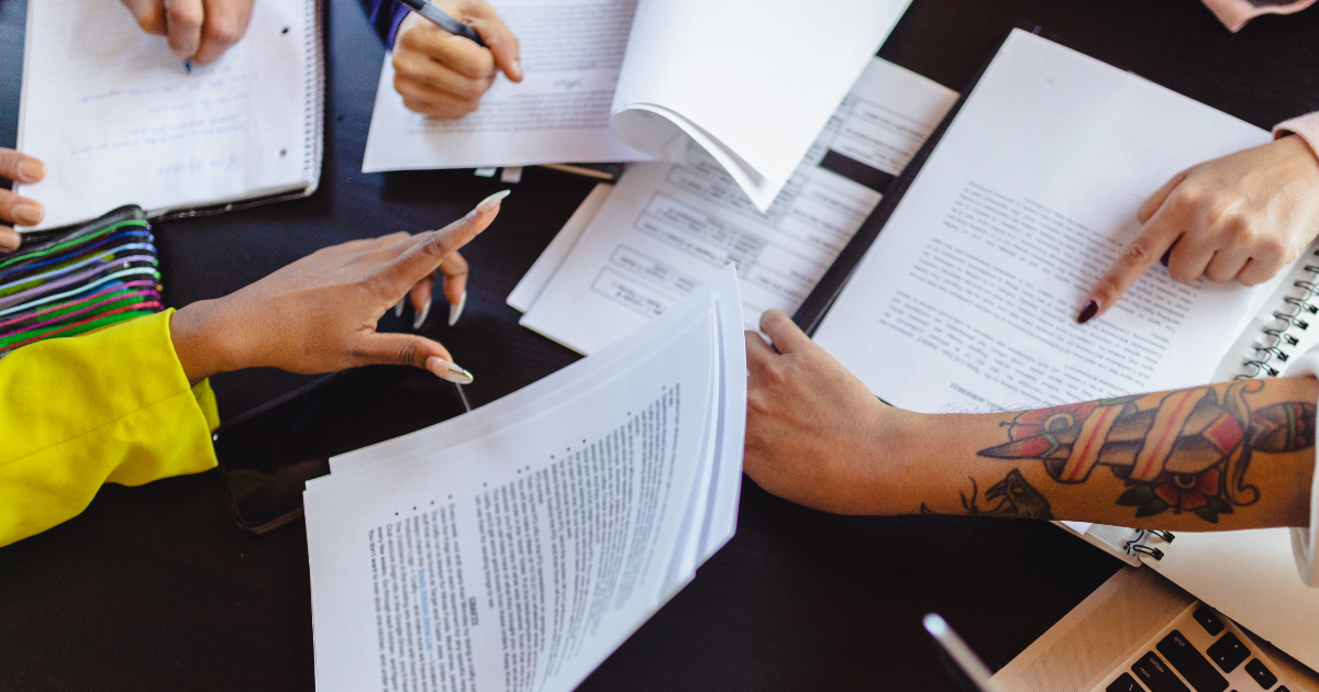 People collaborating at a table with documents, notebooks, and a laptop. Hands are pointing and writing, with one arm showing a tattoo.