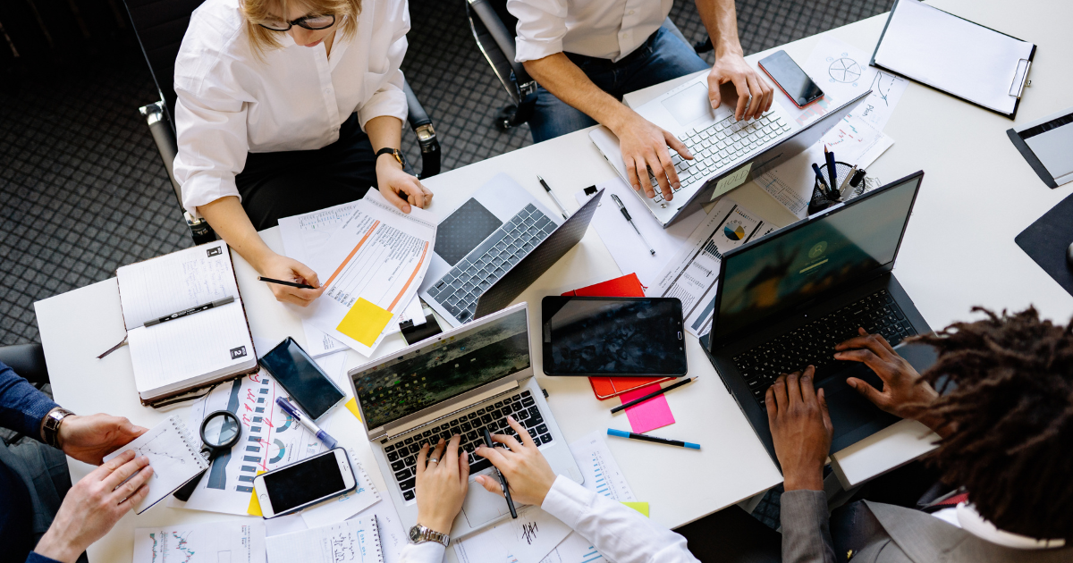 People working collaboratively at a table with laptops, notebooks, and documents, in a busy office setting.