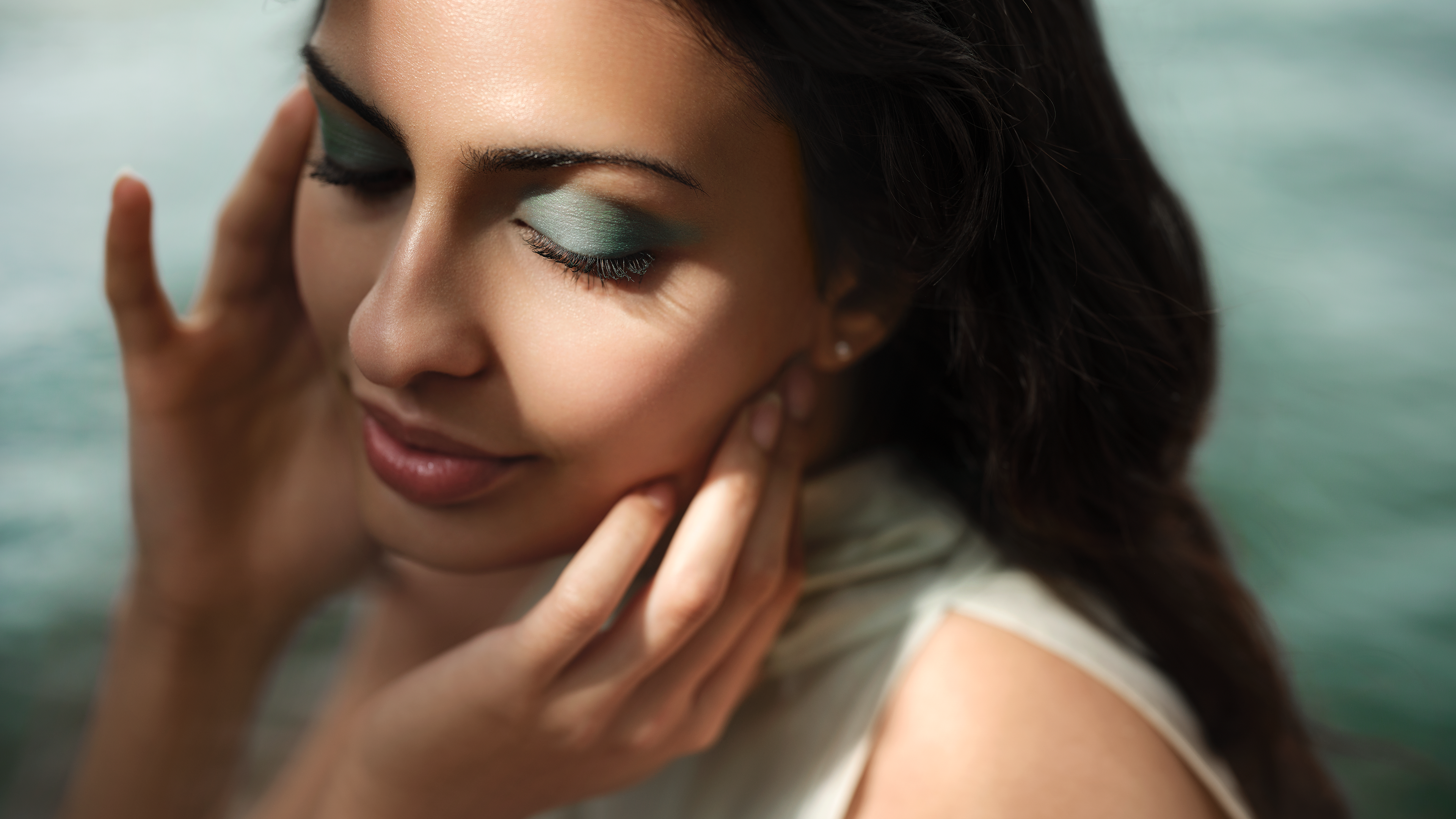 Woman with closed eyes and green eyeshadow, gently touching her face, bathed in soft sunlight with a blurred green background.