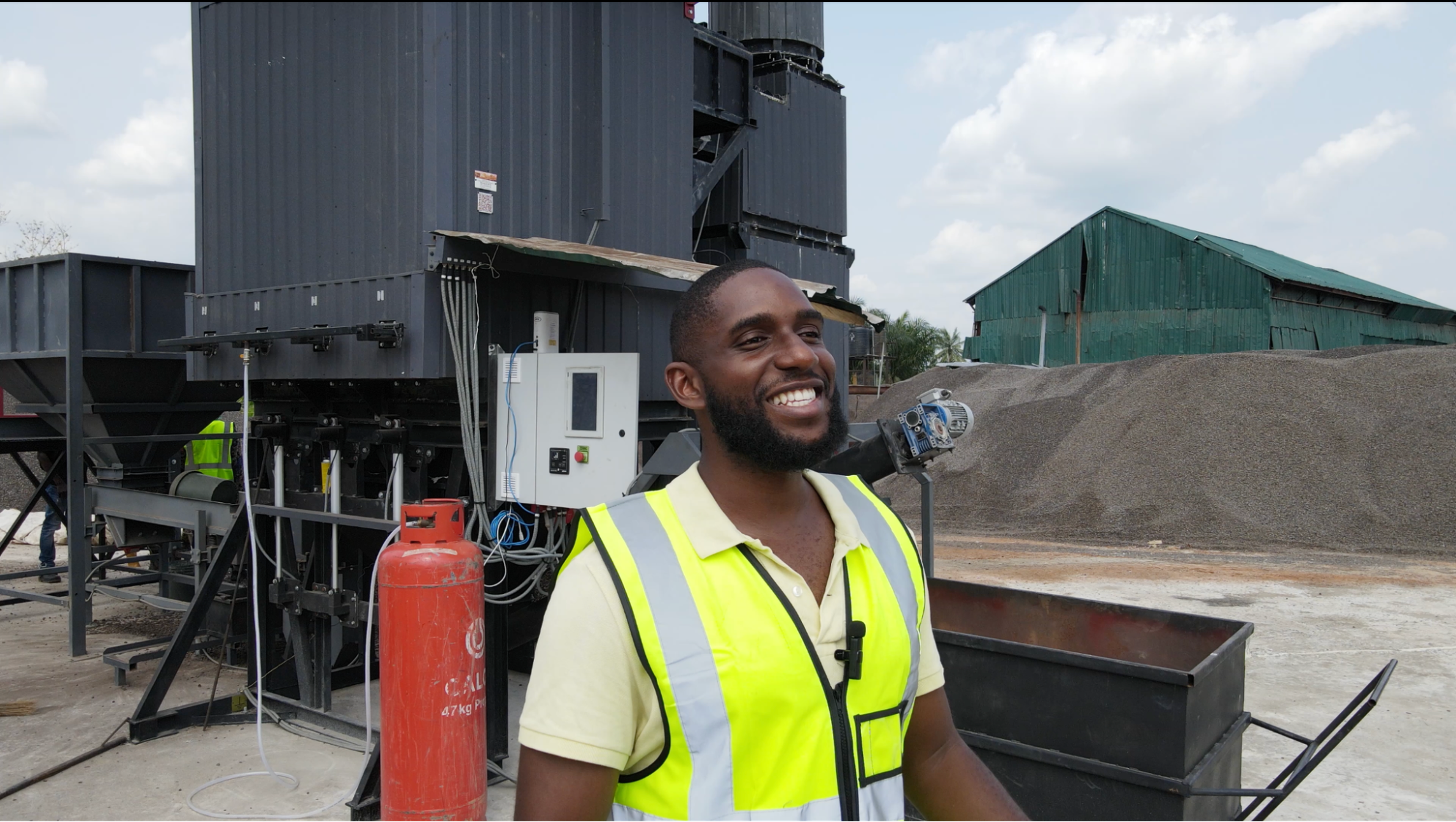 Uzo in a yellow safety vest stands at an industrial site with machinery and piles of gravel in the background.
