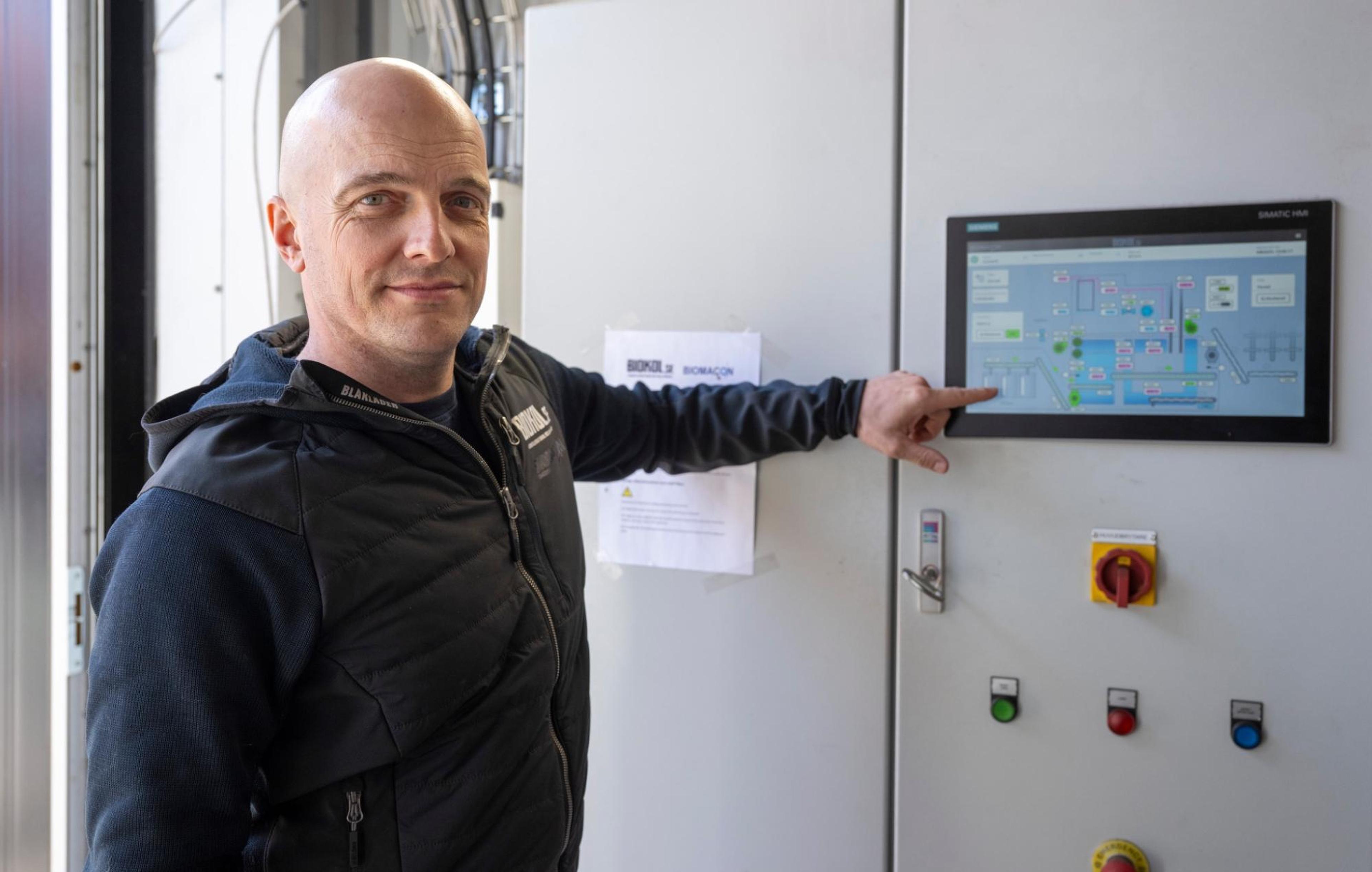 Edvard in a black jacket points at a touchscreen interface on a control panel, displaying a colorful diagram or layout.