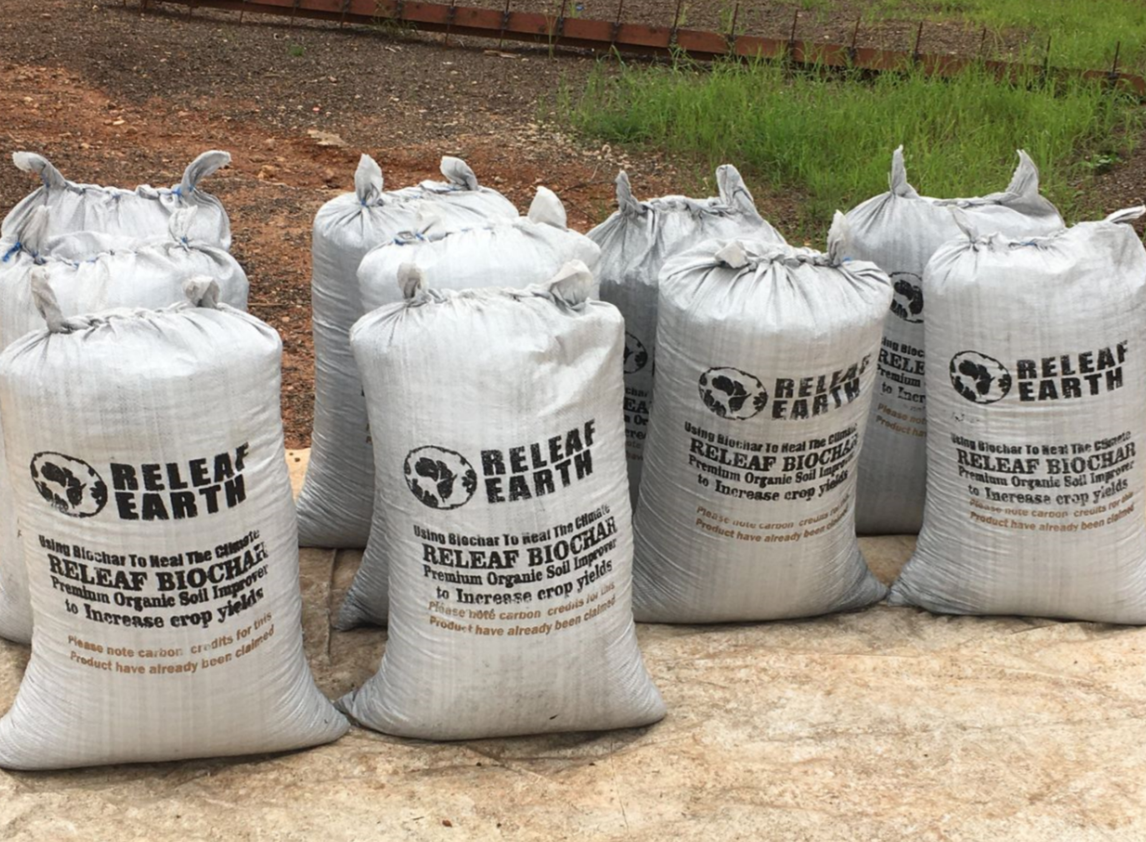 Several bags labeled "Releaf Biochar" by Releaf Earth, stacked outdoors on a concrete surface, advertise organic soil enhancer.