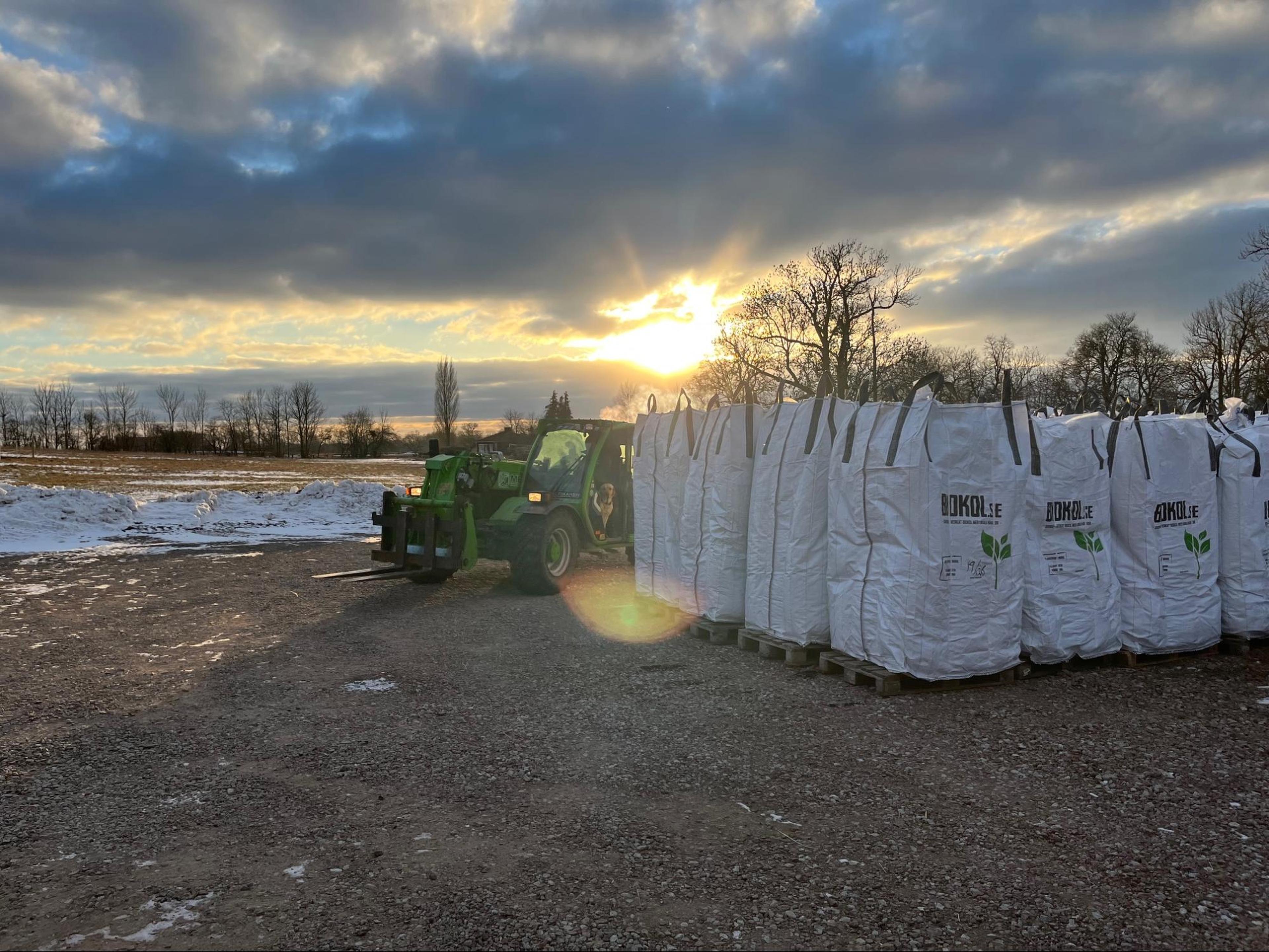 Tractor parked near stacked white bags under a partly cloudy sky, with a setting sun in the background and snow patches on the ground.