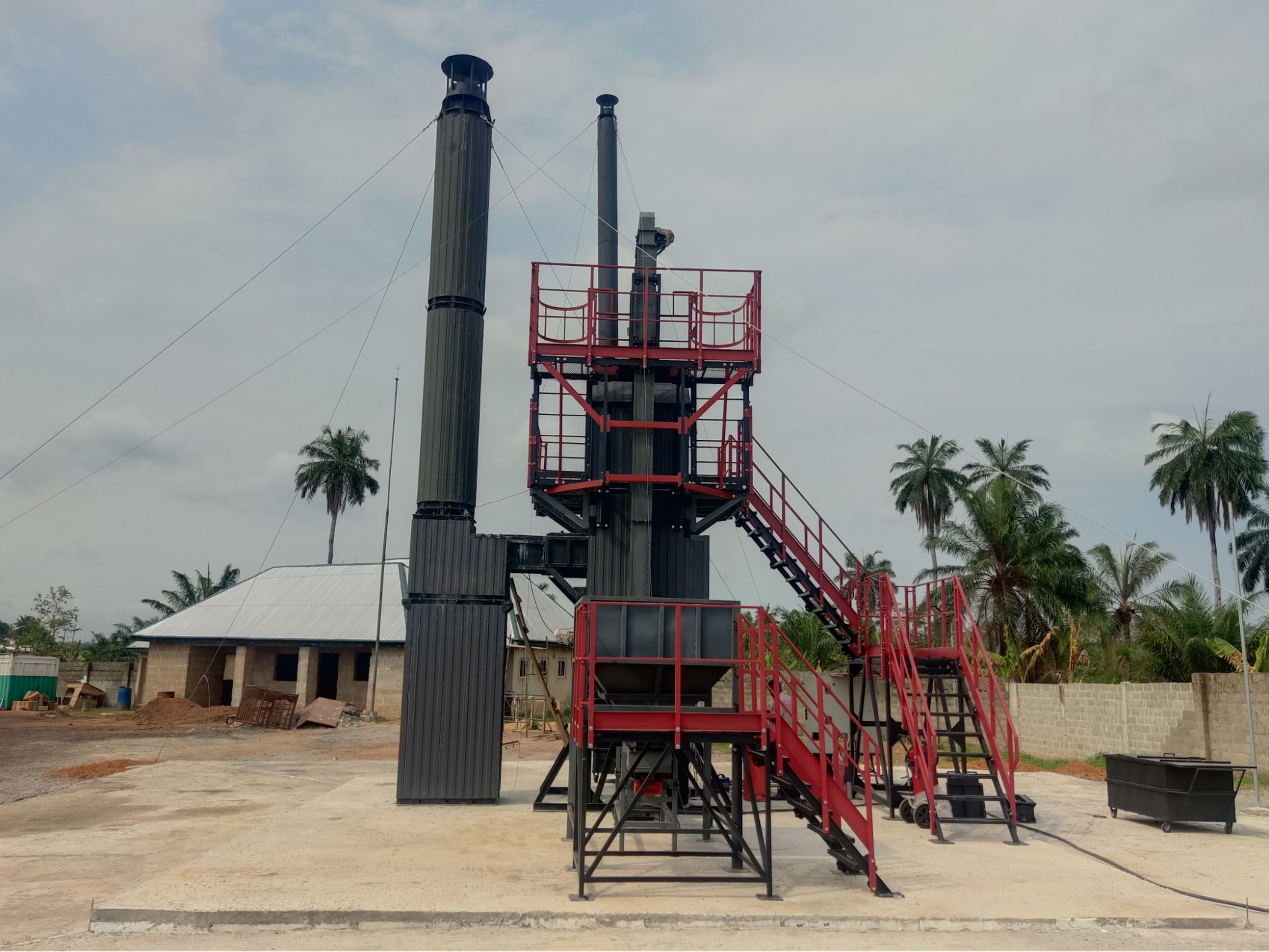 Industrial incinerator with tall chimneys and red staircases outdoors, surrounded by palm trees and a building in the background.