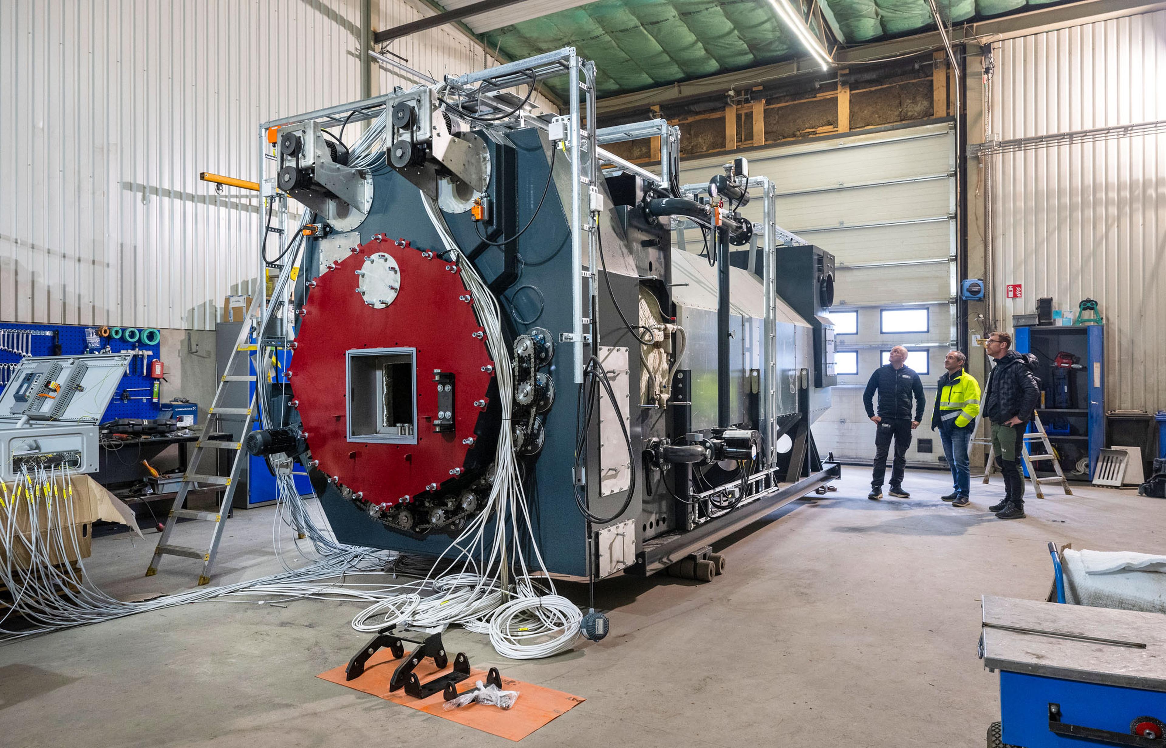 Three people stand in a workshop, examining a large industrial machine with a red front panel and numerous cables.