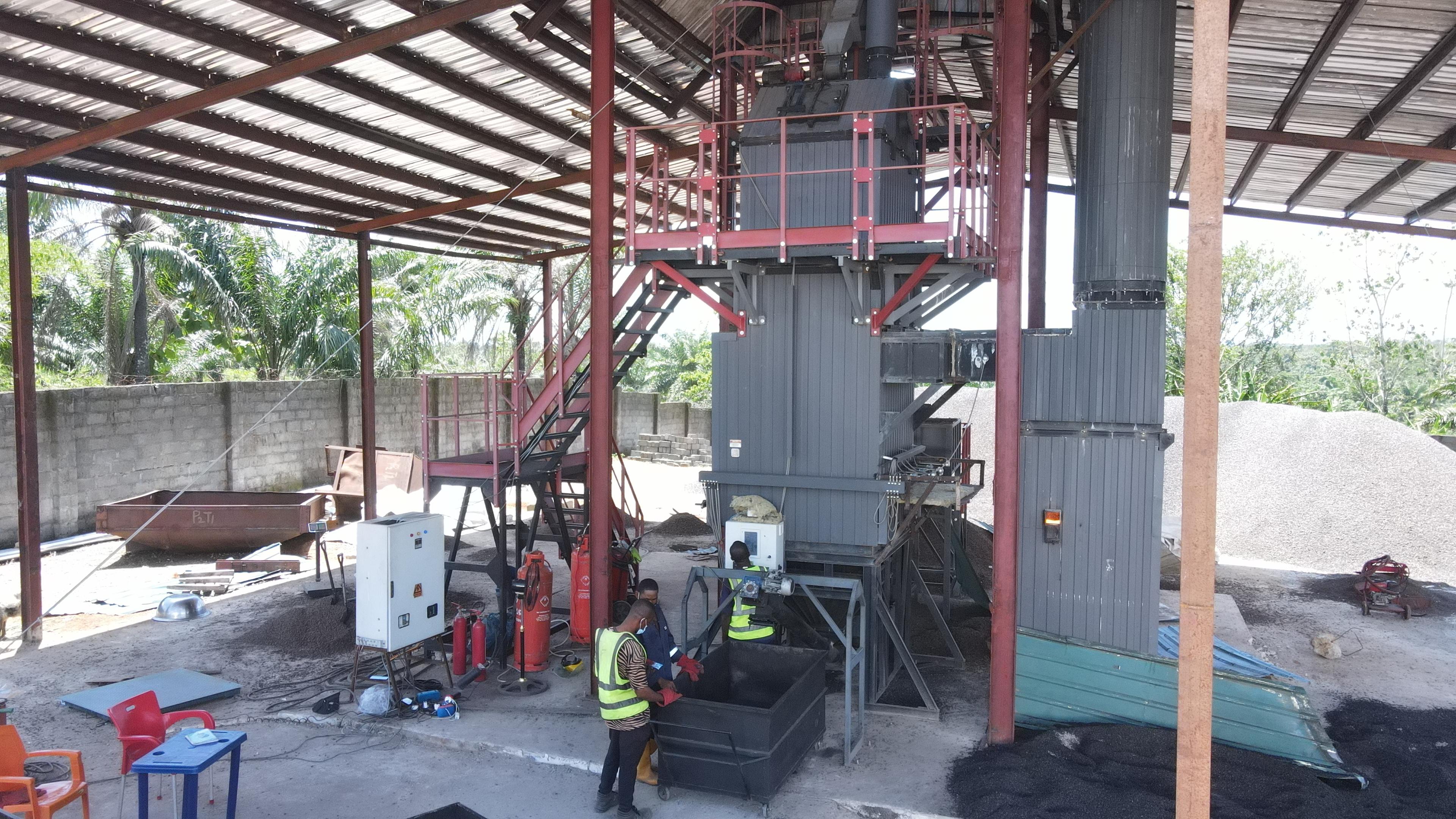An industrial facility with large machinery, workers in safety vests, and heaps of material, under a metal roof, surrounded by greenery.