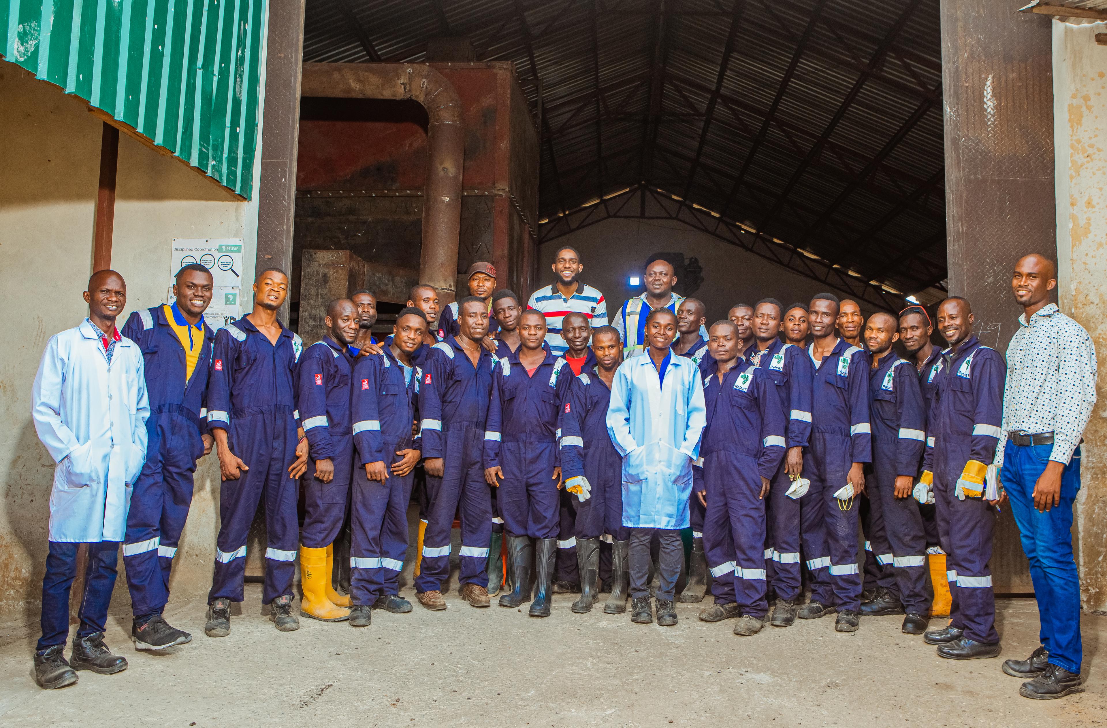 A group of workers in blue uniforms and white coats pose together in an industrial setting with a large open entrance behind them.