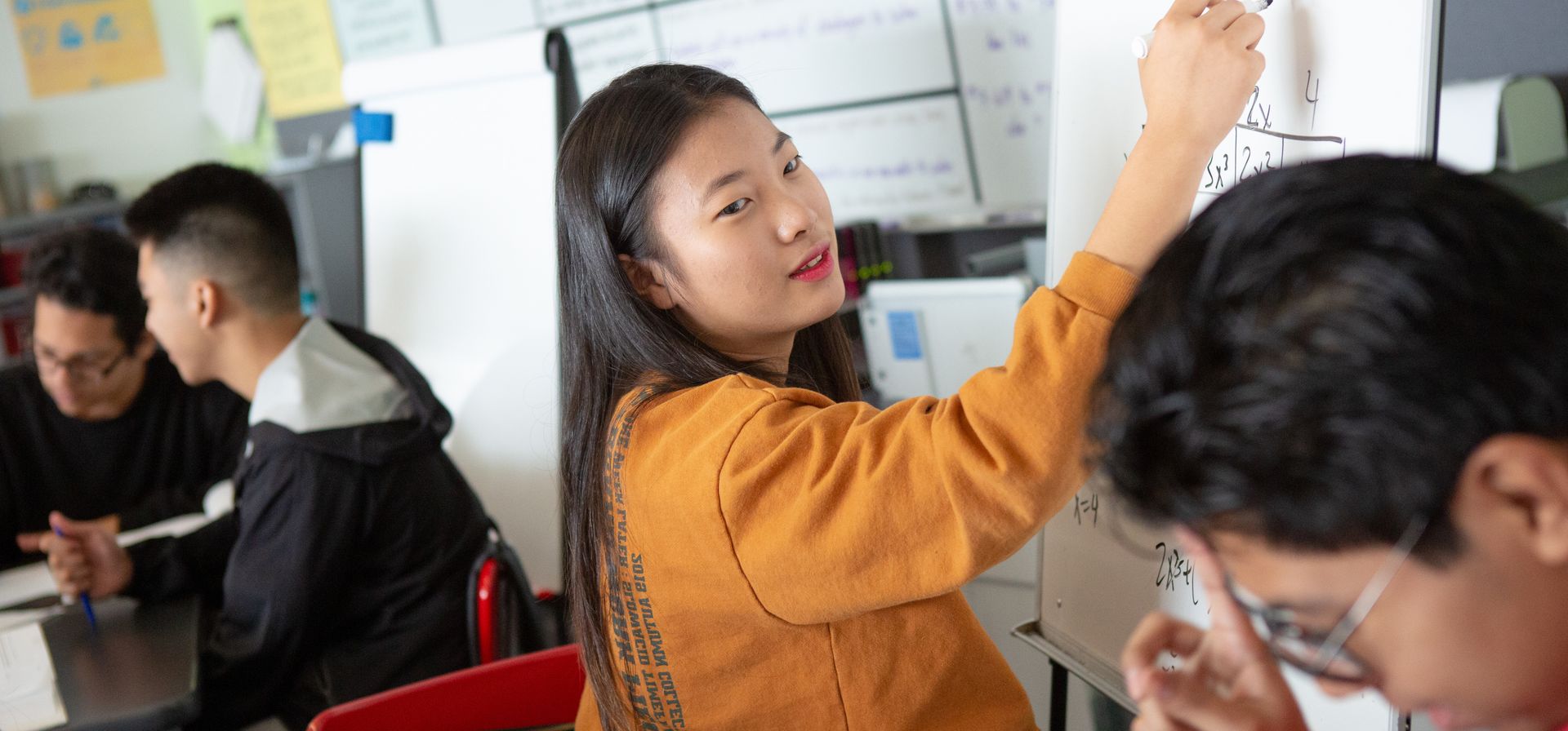 A student in an orange sweater writes on a whiteboard while others sit at desks, collaborating in a classroom setting.