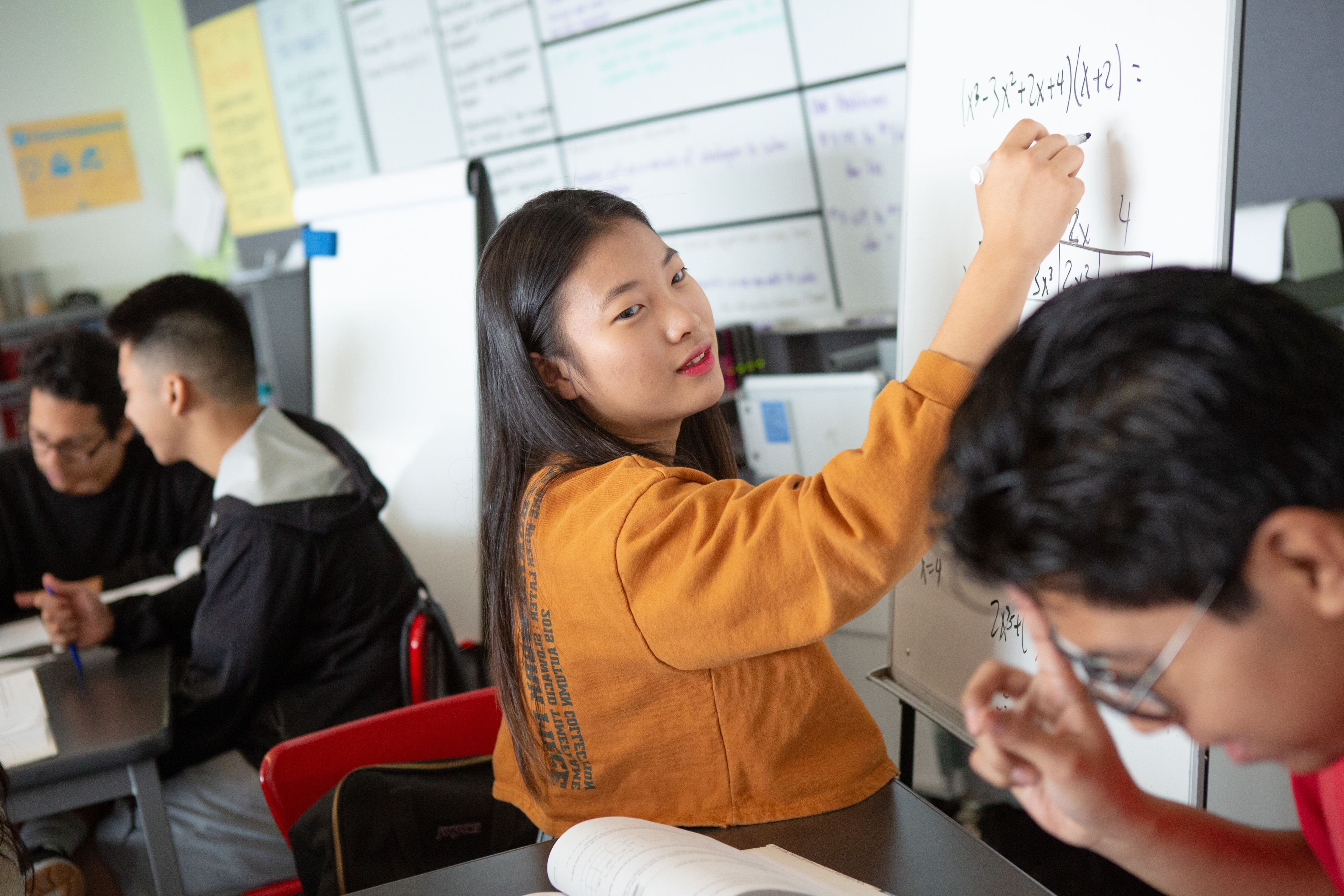 A student in an orange sweater writes on a whiteboard while others sit at desks, collaborating in a classroom setting.