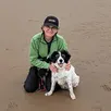 Person in a green jacket and cap kneels on a sandy beach, smiling while holding a black and white dog.