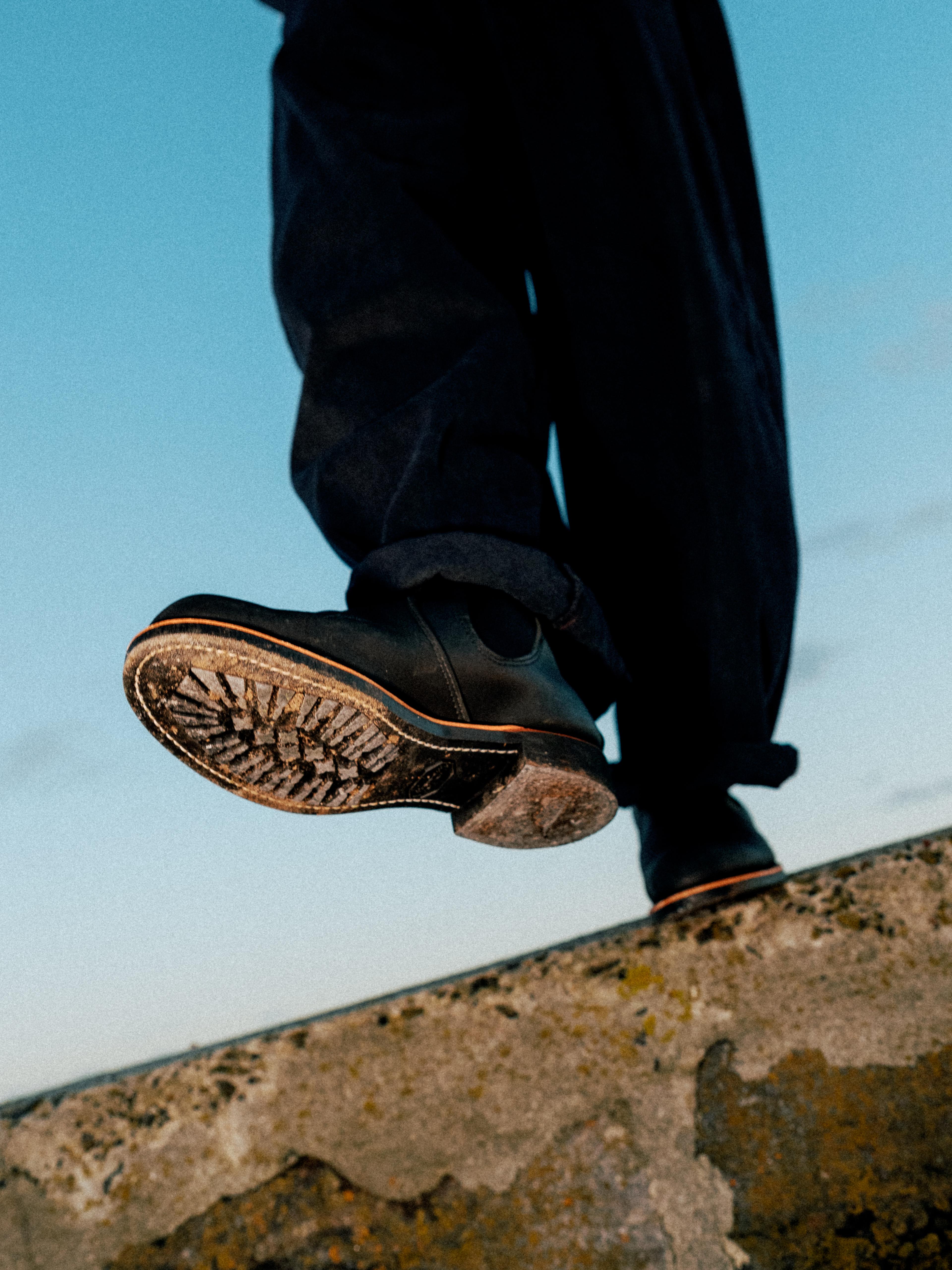 Person stepping up on a ledge, wearing dark pants and black shoes, with a textured sole. Background features a clear blue sky.