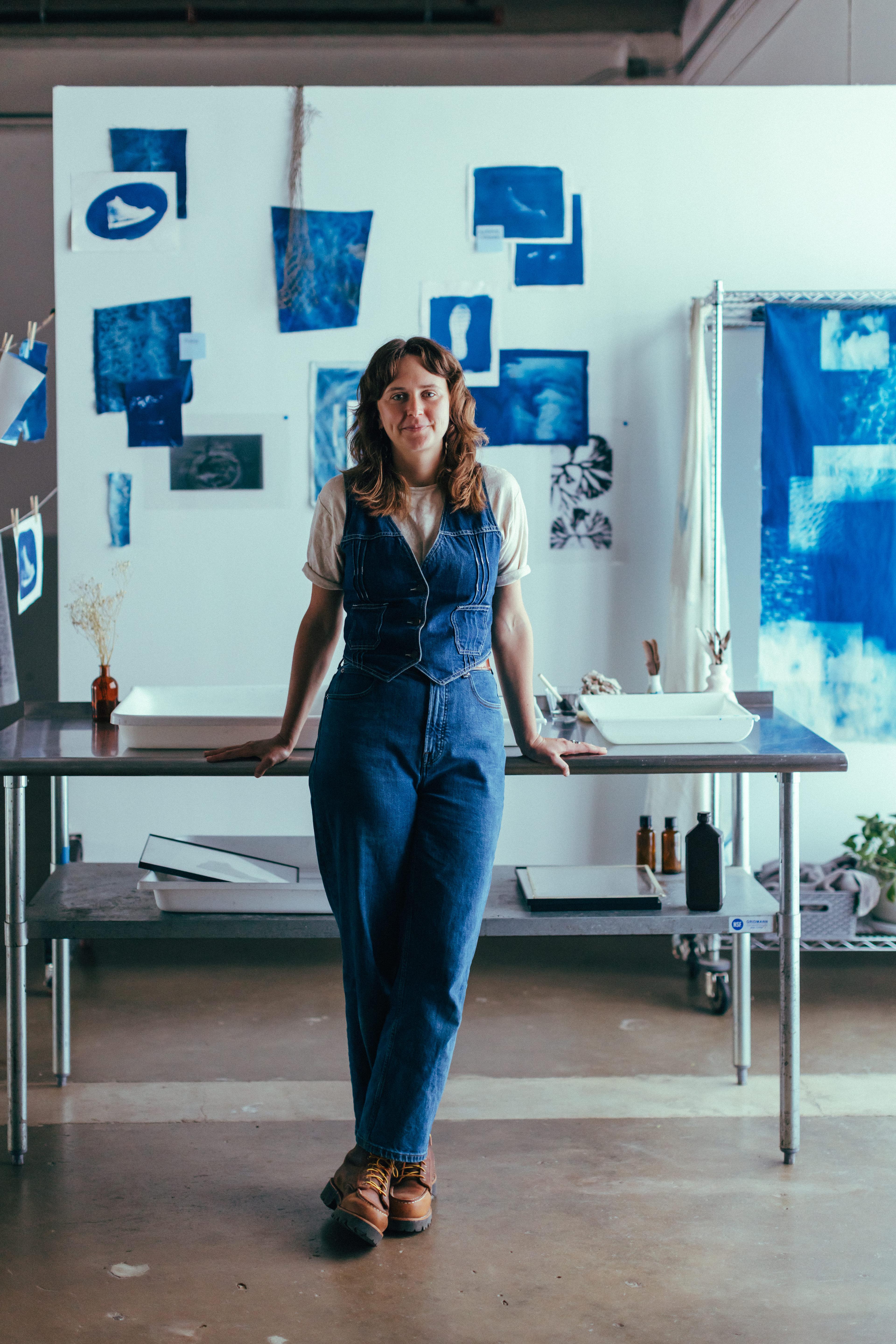 Person standing in an art studio, leaning against a table, with blue artworks displayed on a white wall behind them.