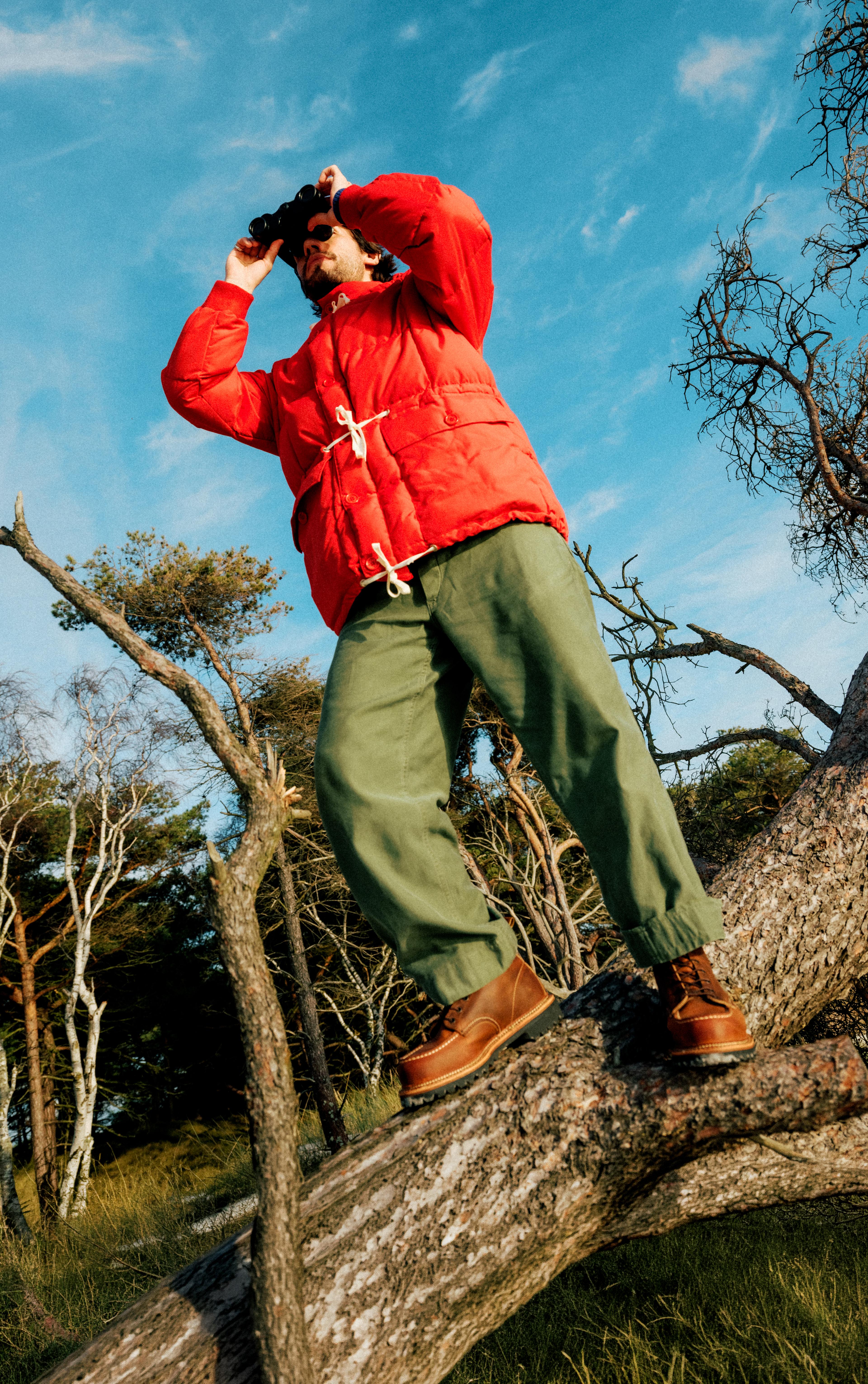 A peron with a red jacket with a Red Wing Boot - standing on a branch looking onwards with binoculars
