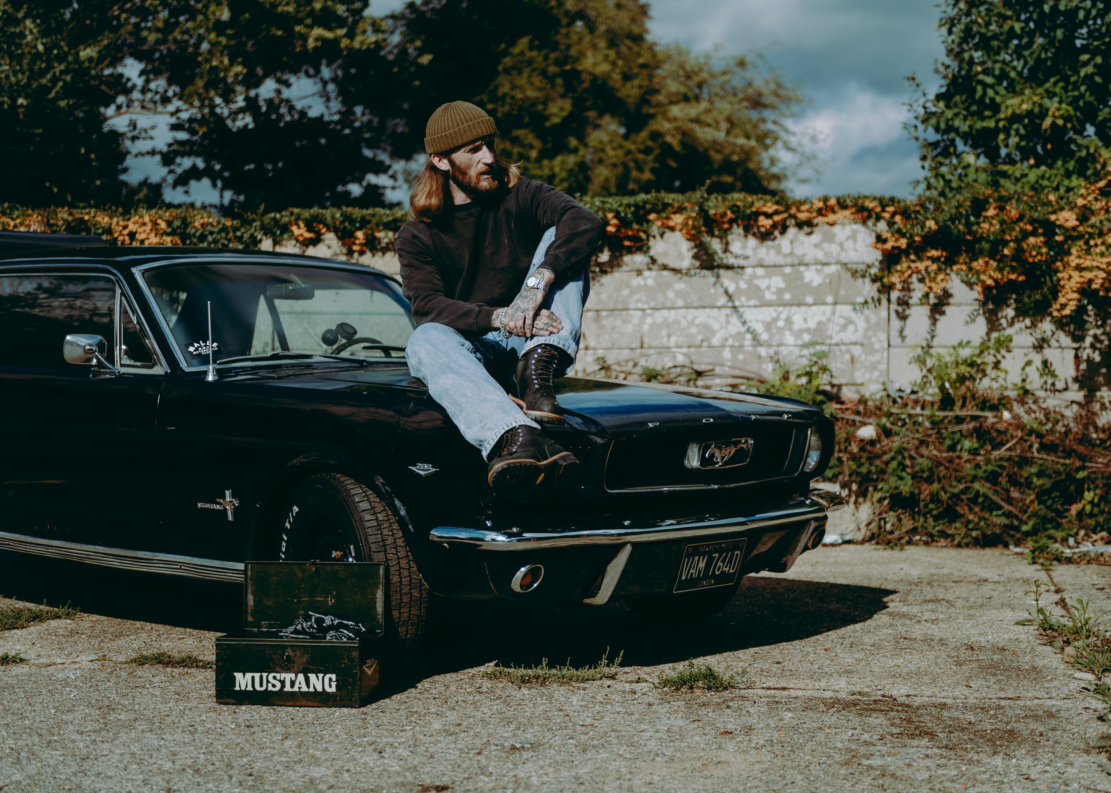 A person in casual attire sits on the hood of a vintage Ford Mustang with a "Mustang" toolbox nearby, against a leafy backdrop.