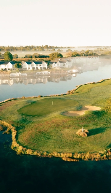 Aerial view of a golf course by a lake, with a small sand bunker. Houses and trees line the foggy waterfront in the background.