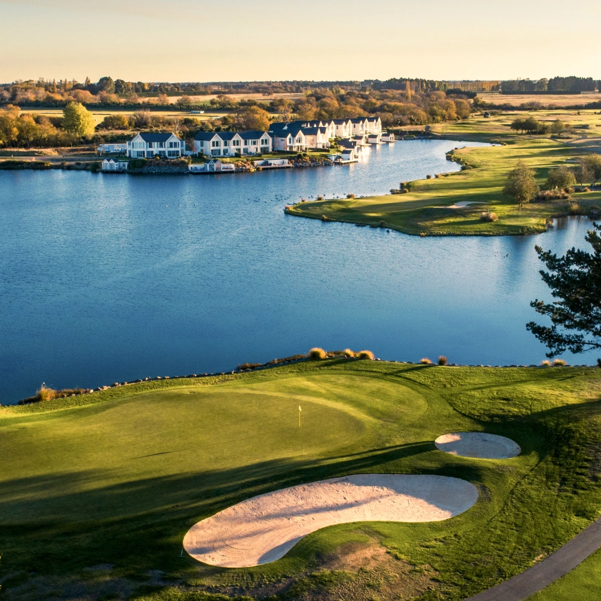 Scenic view of a golf course by a lake at sunset, with sand traps, a tree, and houses along the shoreline in the background.