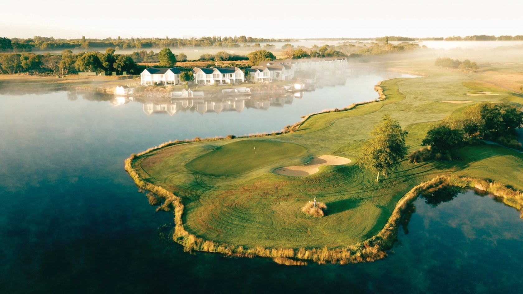 Aerial view of a serene golf course by a lake at sunrise, with mist hovering over the water and houses in the background.
