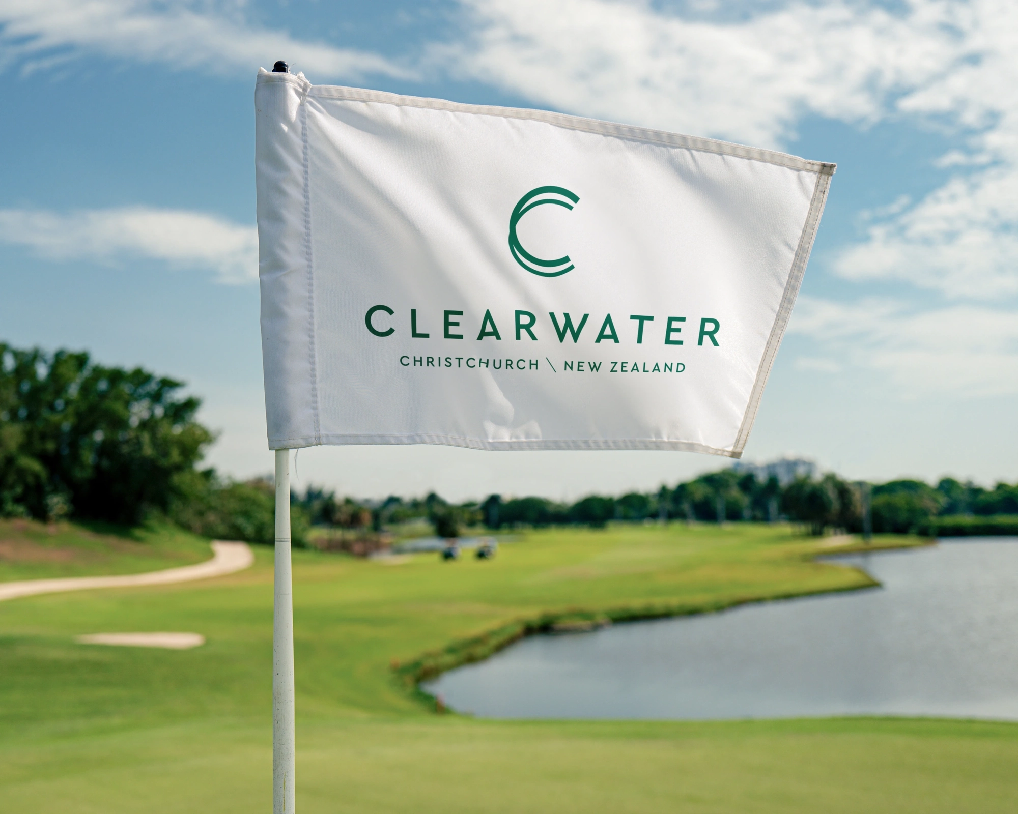 Golf course with a "Clearwater, Christchurch, New Zealand" flag. Lush greenery, a water hazard, and a cloudy sky in the background.