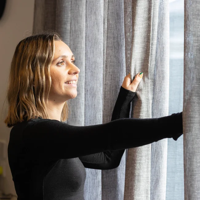 Smiling woman drawing back sheer grey linen curtains at a window