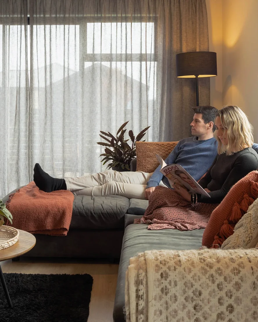 Couple relaxing on a sofa beside sheer grey curtains in a warm living room