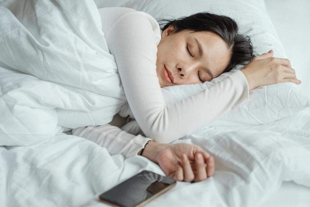 Woman sleeping peacefully in bed, wearing a white long-sleeve shirt, with a smartphone resting nearby on the white sheets.