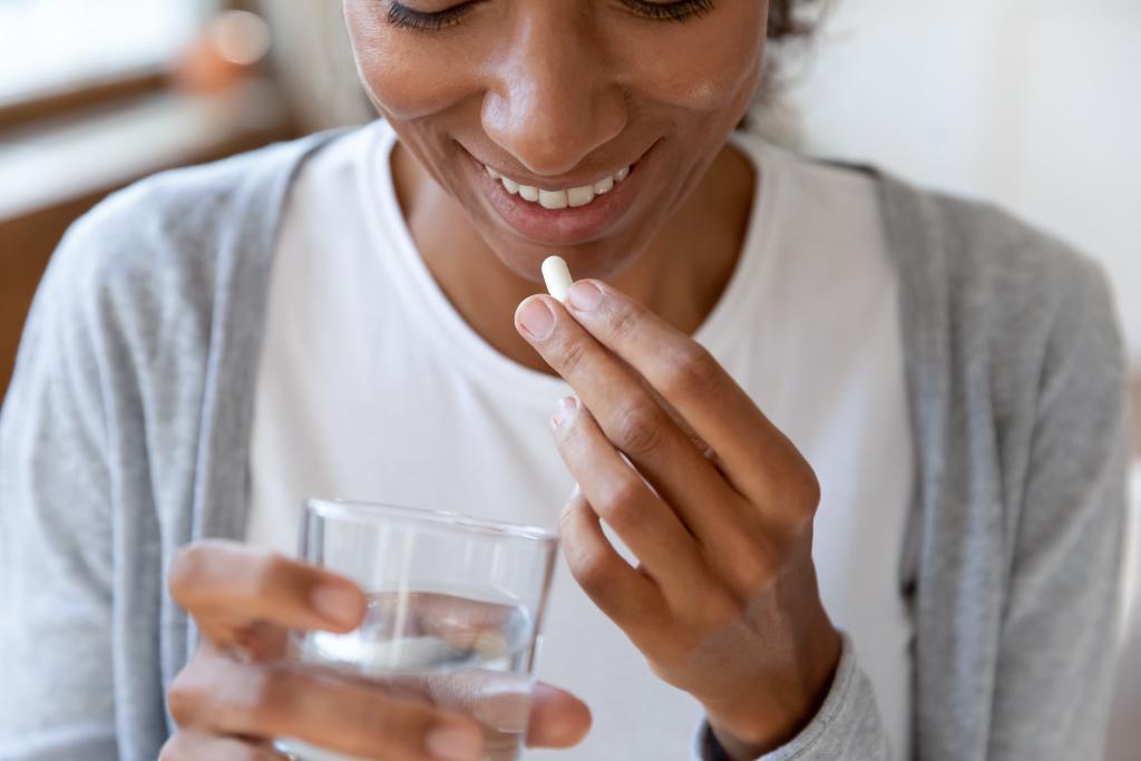 A person smiling, holding a pill in one hand and a glass of water in the other, preparing to take the medication.