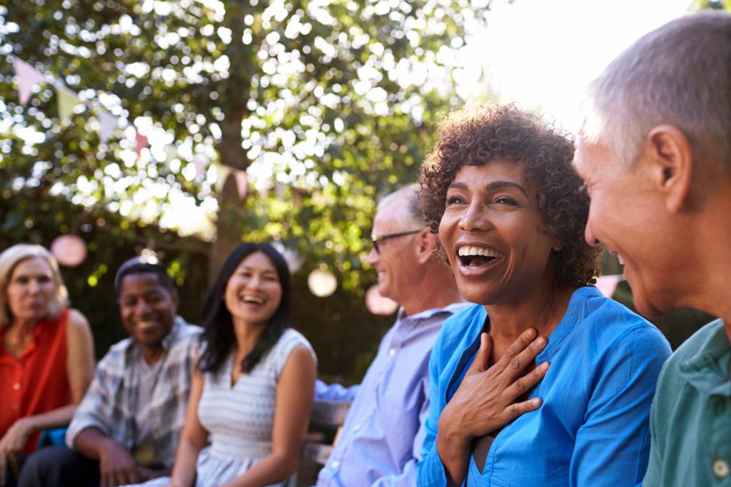 A diverse group of people sitting outdoors, smiling and laughing together, with trees and string lights in the background.