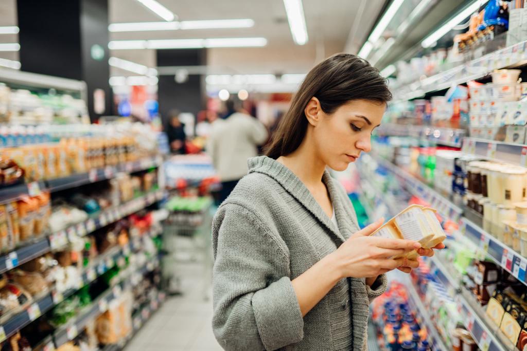 Woman reading a product label in a grocery store aisle, surrounded by shelves stocked with various items.