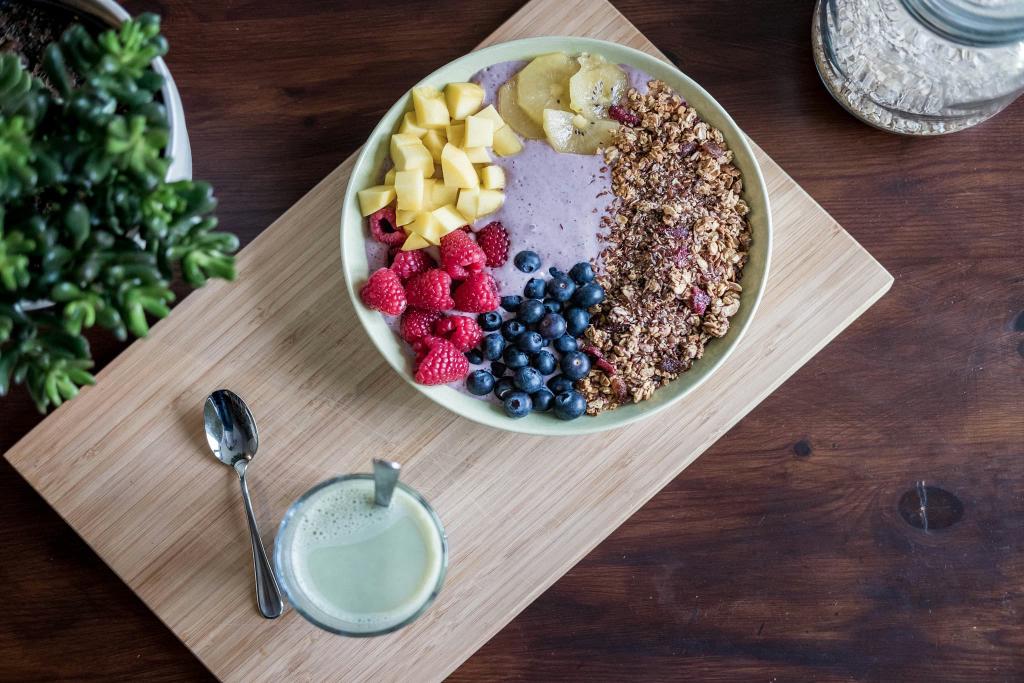 A smoothie bowl with granola, mango, raspberries, blueberries, and banana on a wooden board, next to a spoon and a green drink.