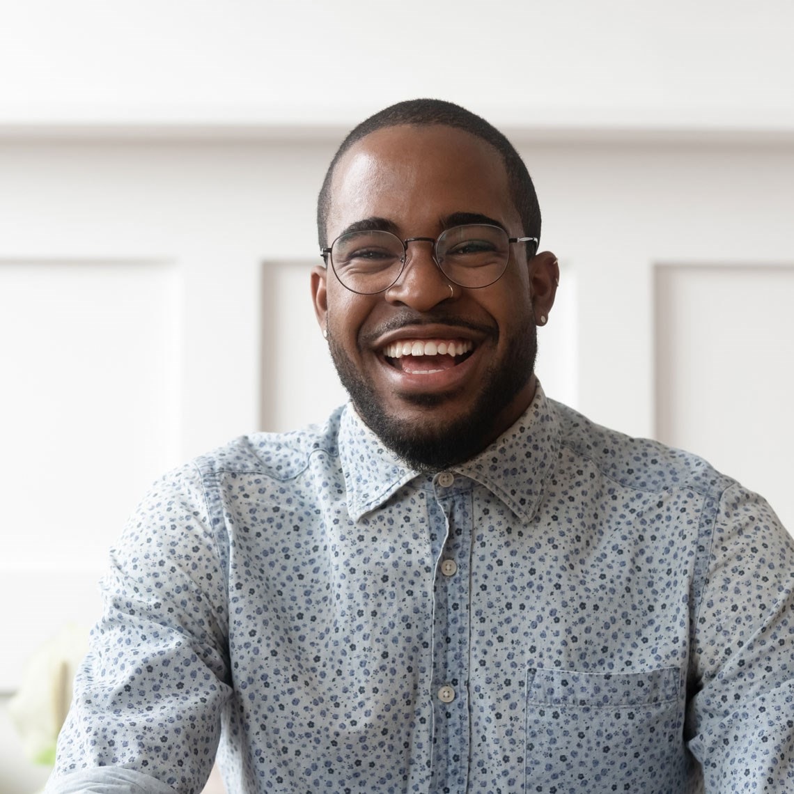 Smiling man with glasses and a beard wearing a patterned button-up shirt, sitting against a white paneled background.