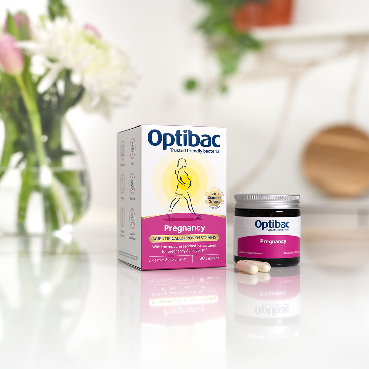 Optibac Pregnancy probiotics box and jar with capsules on a reflective surface, surrounded by blurred flowers in the background.