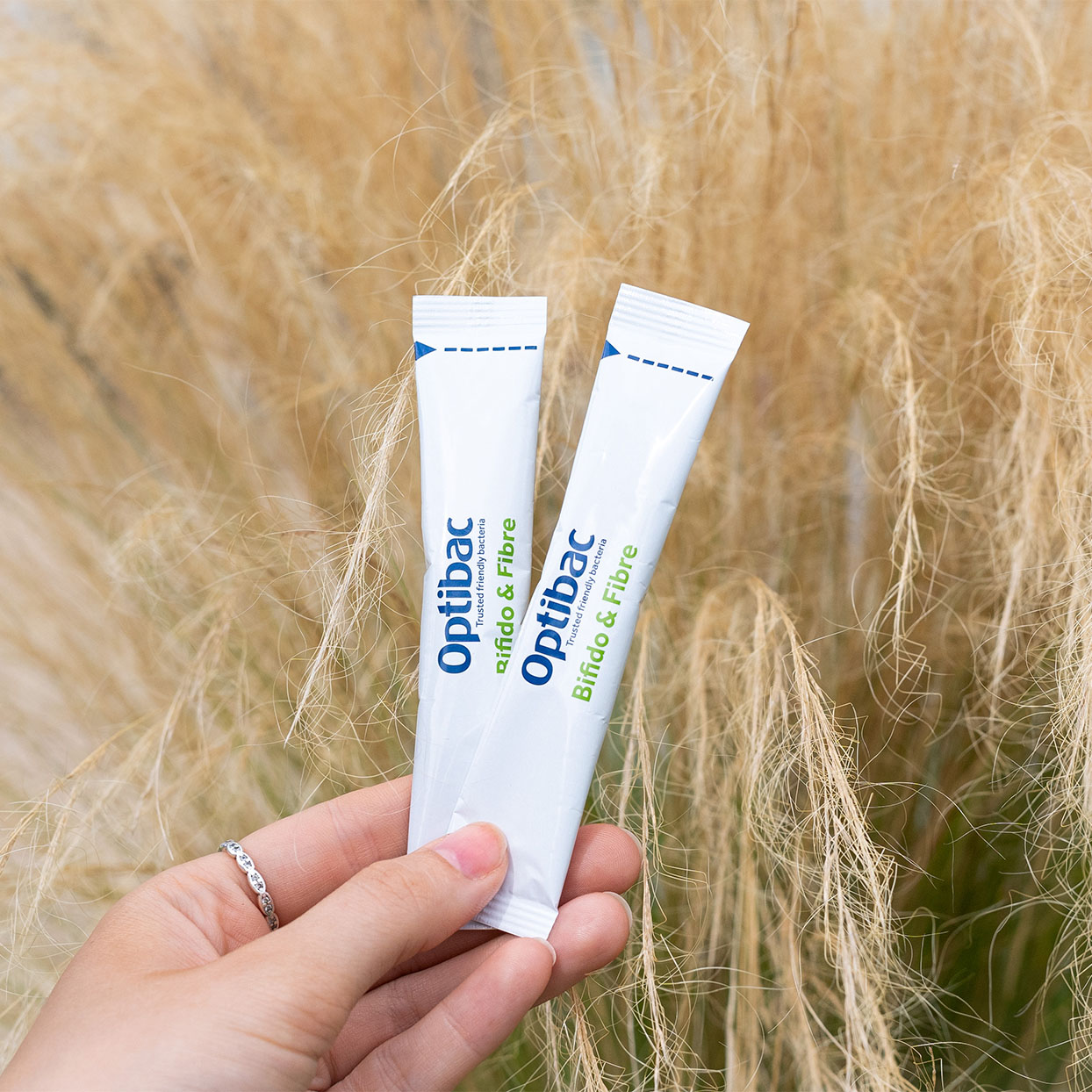 Hand holding two Optibac sachets against a background of tall, beige grass.