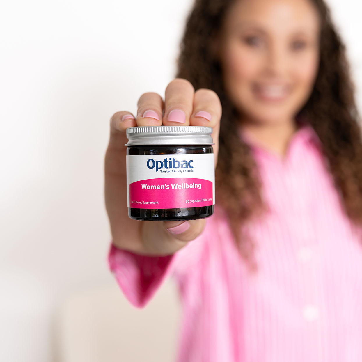 Woman in a pink shirt holding a jar labeled "Optibac Women's Wellbeing" towards the camera, smiling in a bright setting.