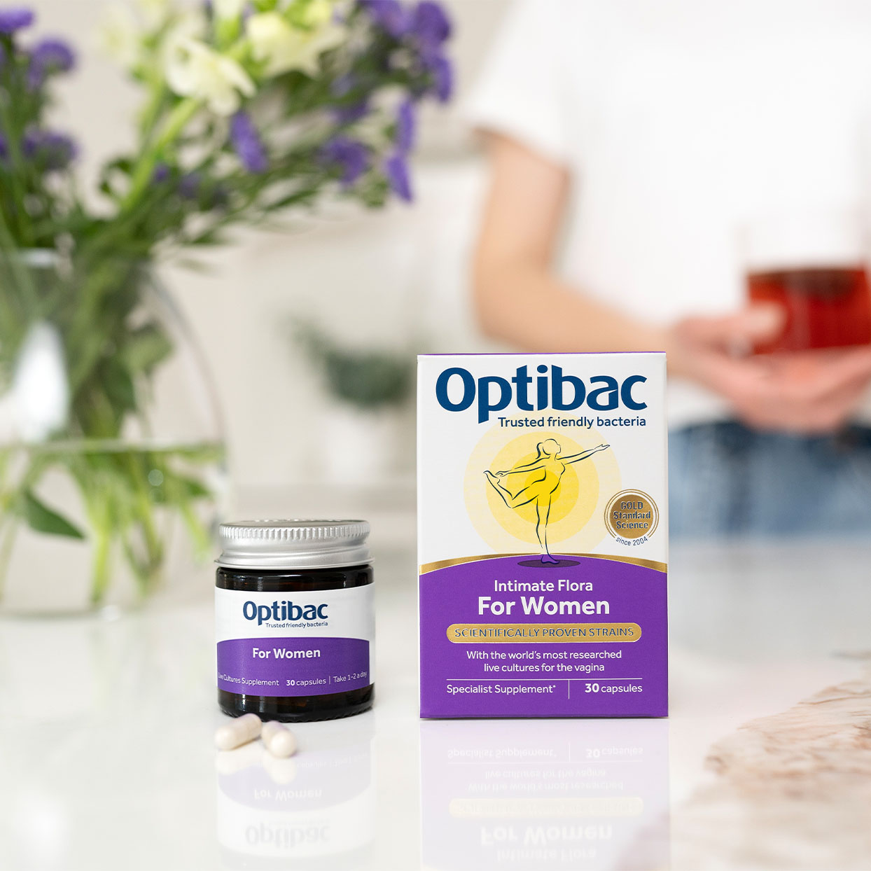 Optibac Intimate Flora for Women on a table with capsules, flowers, and a blurred person in the background.