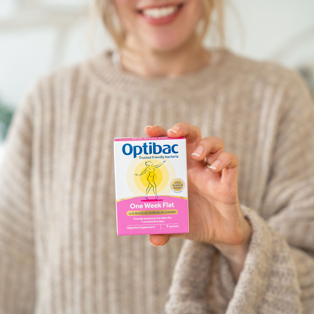 Person holding a box of Optibac One Week Flat probiotics, smiling, wearing a beige sweater.