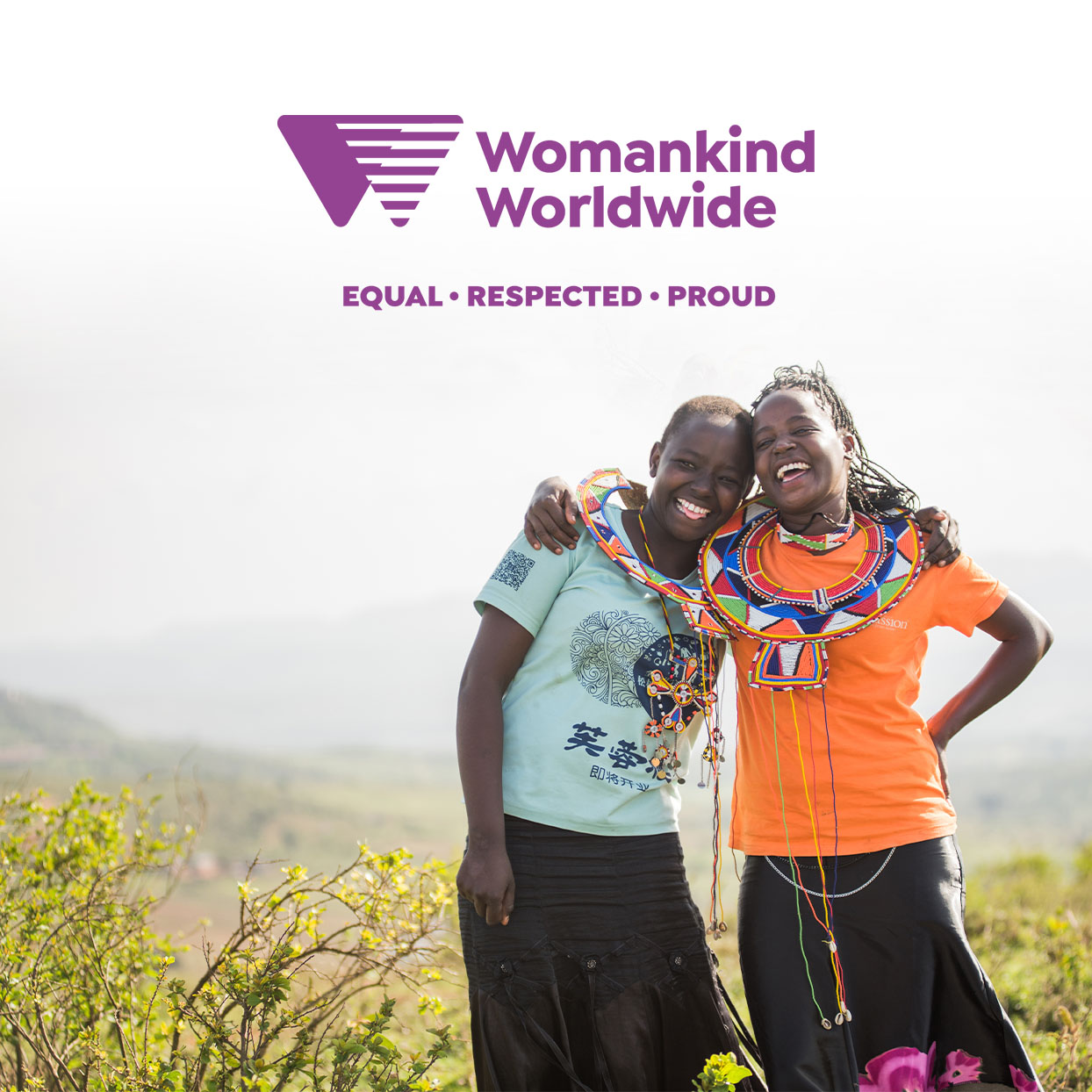 Two smiling women in colorful attire embrace outdoors, with "Womankind Worldwide" and the slogan "Equal • Respected • Proud" above them.