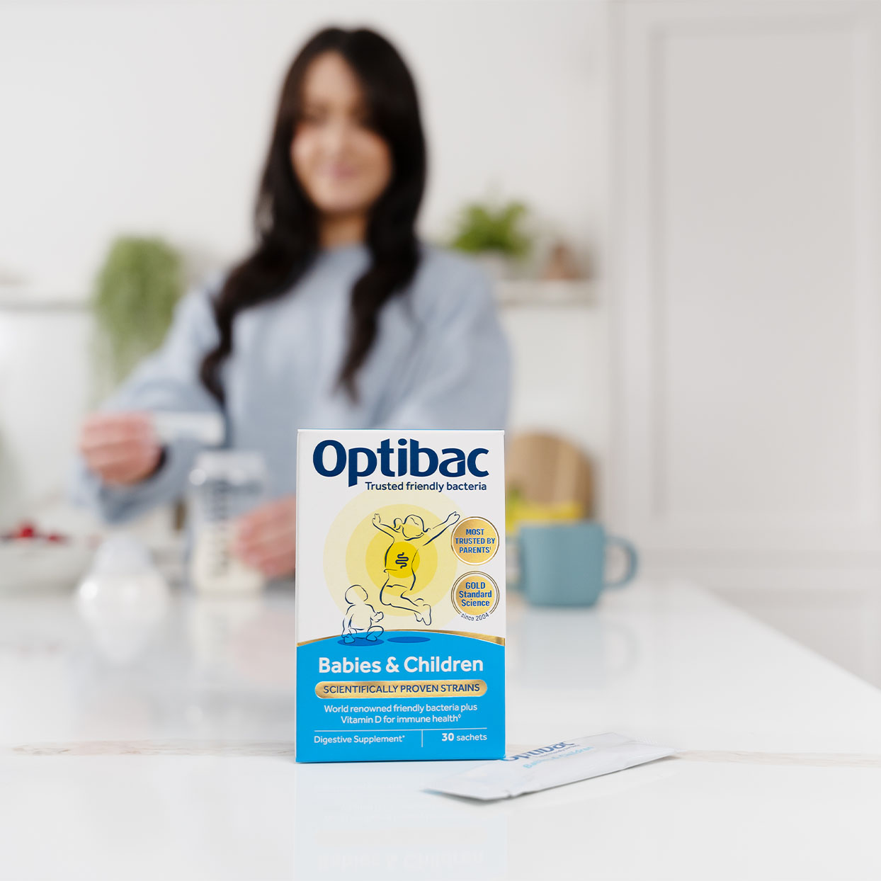 Box of Optibac probiotics for babies and children on a kitchen counter, with a person in the background preparing a drink.