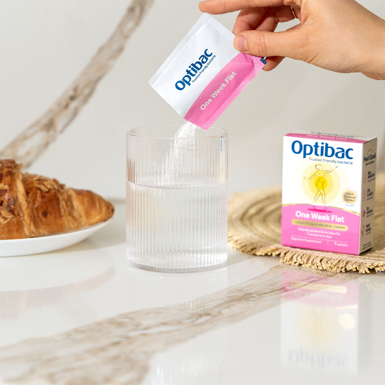 Hand pouring Optibac powder into a glass of water on a table with a croissant and Optibac box nearby.