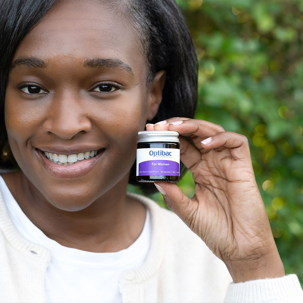 A person smiling and holding a small jar labeled "Optibac For Women" against a blurred green background.