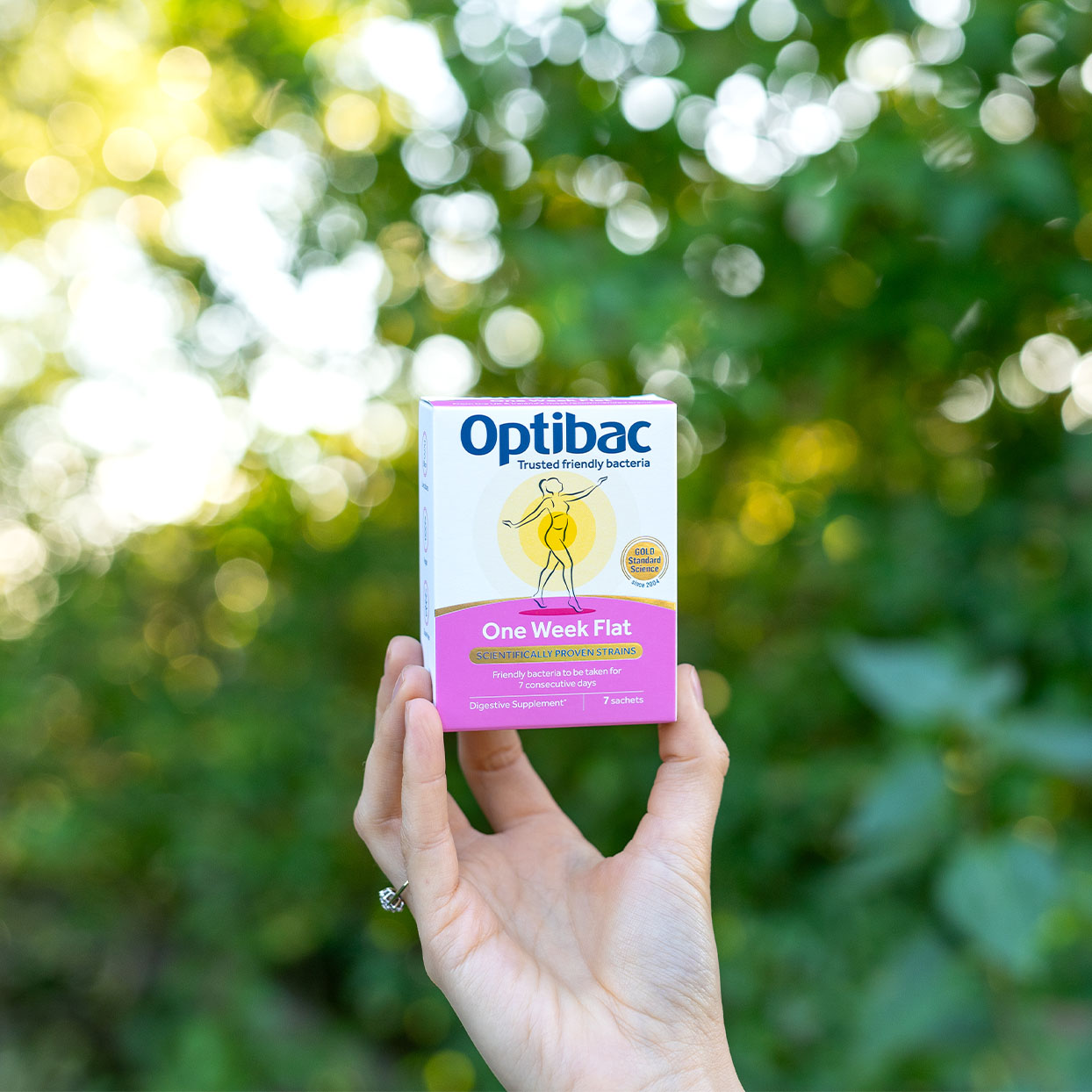 Hand holding an Optibac One Week Flat probiotic box against a blurred green leafy background.