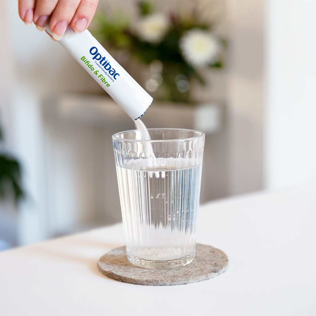 A hand pours Optibac probiotic powder into a glass of water on a coaster, with blurred flowers in the background.