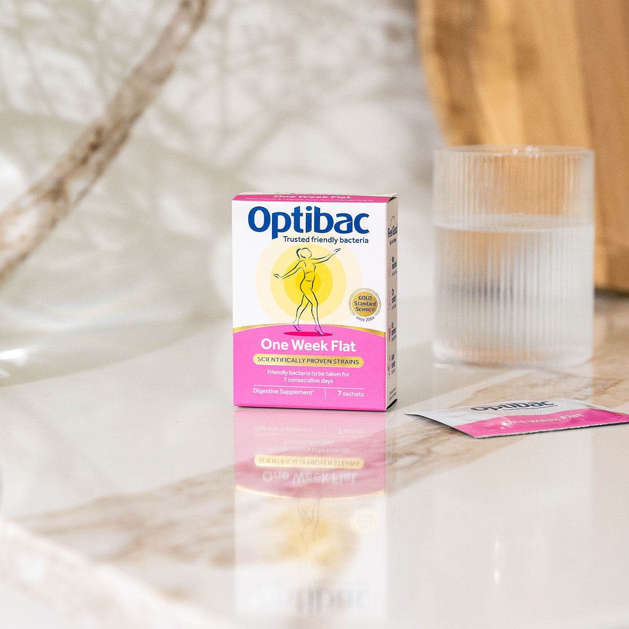 Optibac One Week Flat probiotic box on a marble surface, with a glass of water and a sachet nearby.