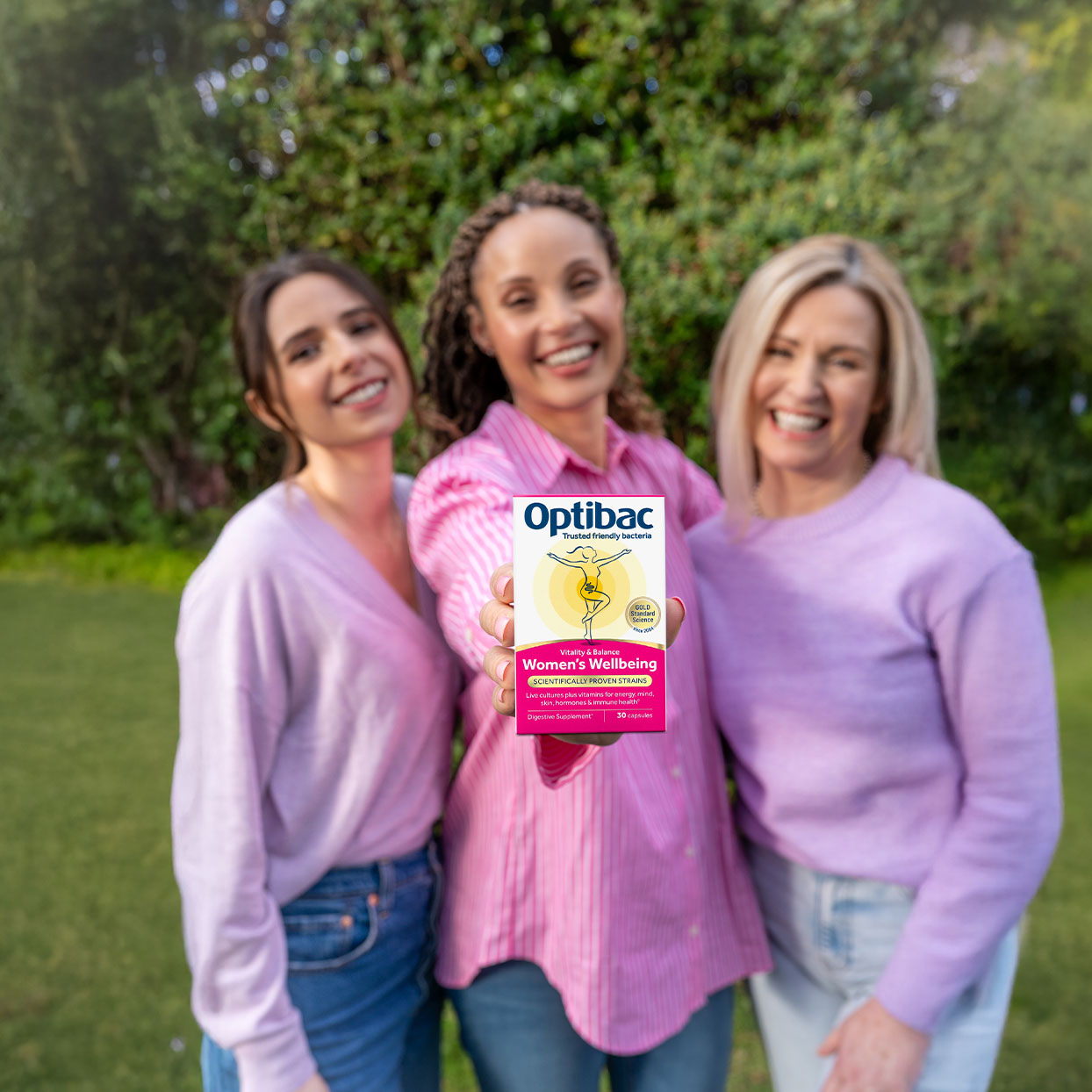 Three women smiling outdoors, holding a box of Optibac Women's Wellbeing supplements. They're dressed in casual pink and purple tops.
